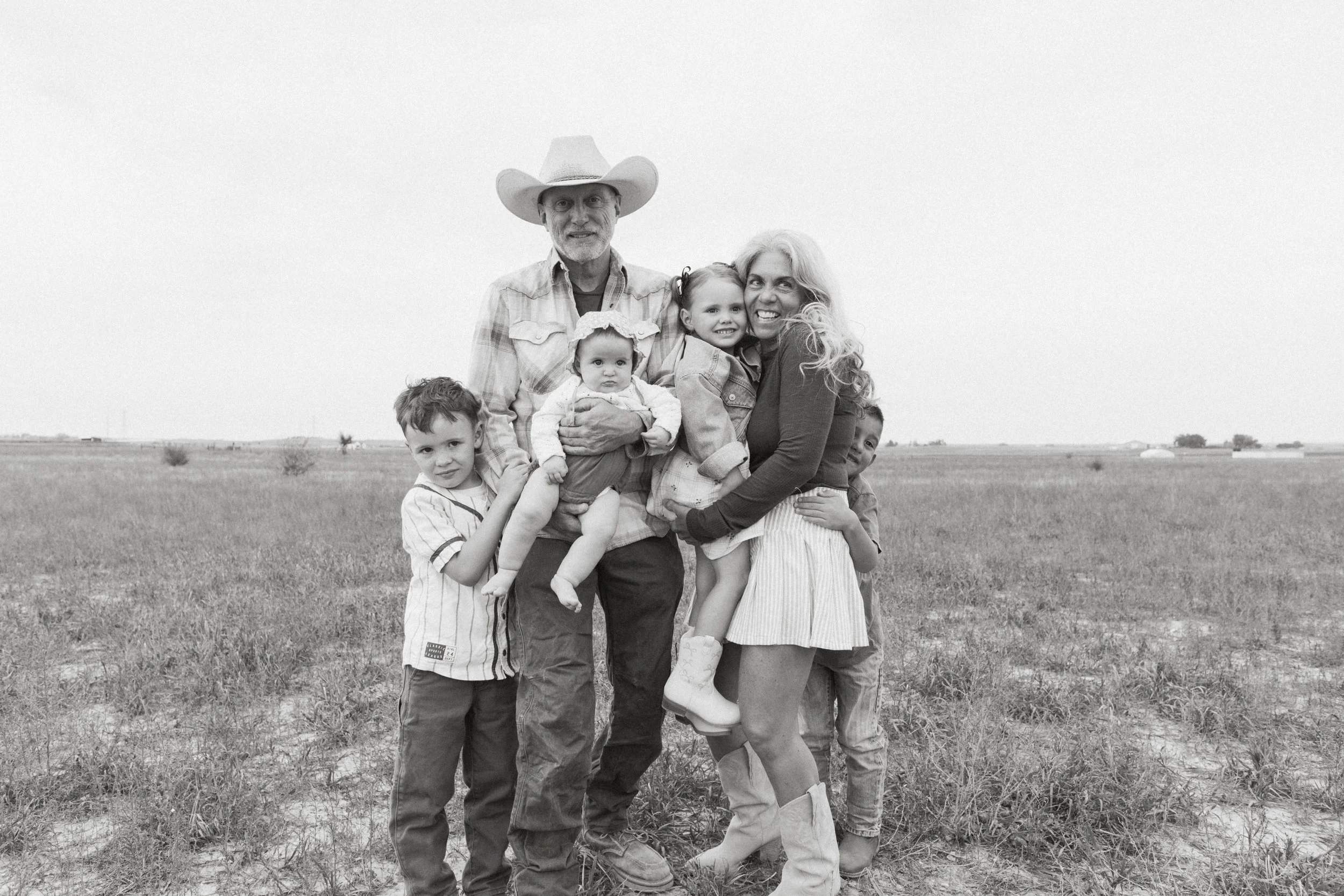 A family of six standing in an open field. The family includes an older man wearing a cowboy hat, an older woman, two young girls, and two young boys. They are smiling and embracing each other.