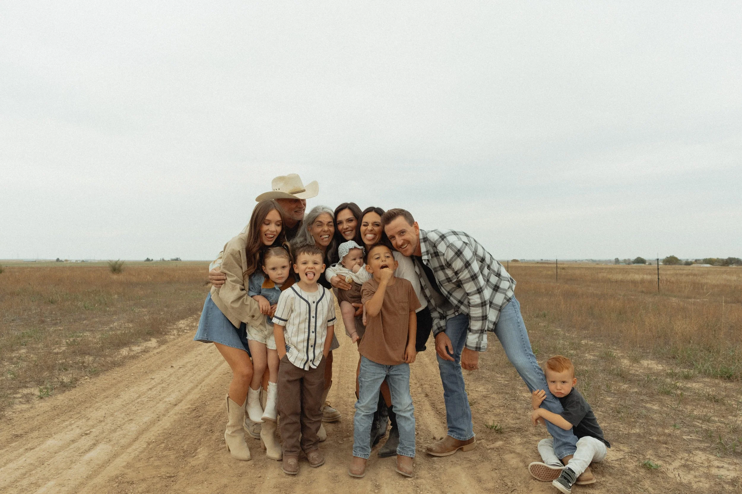 A group of people, including children and adults, standing on a dirt road in an open field under a cloudy sky, posing for a photo and smiling.