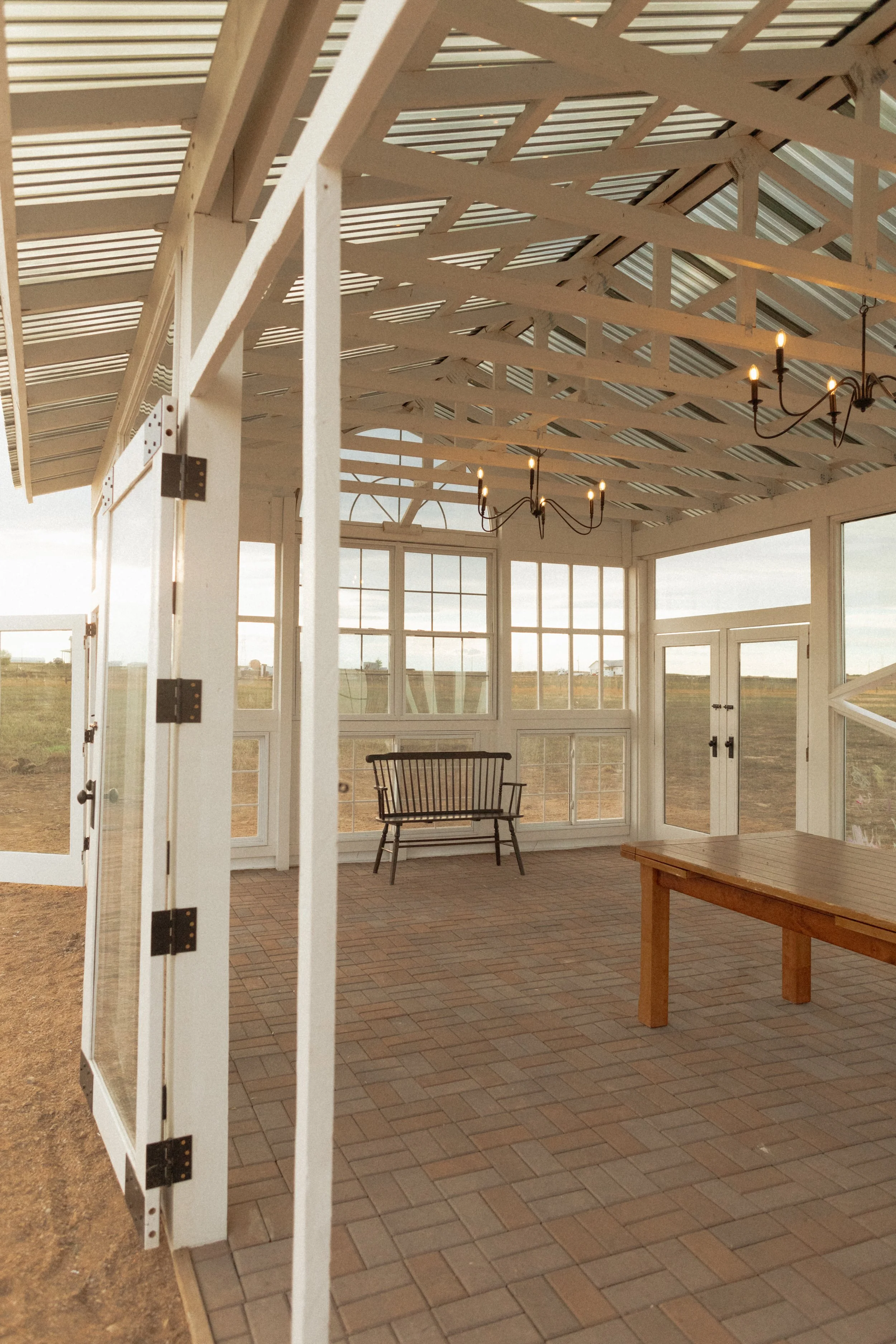 Sunlit conservatory with brick floor, wooden bench, chandelier, and large windows overlooking open land.