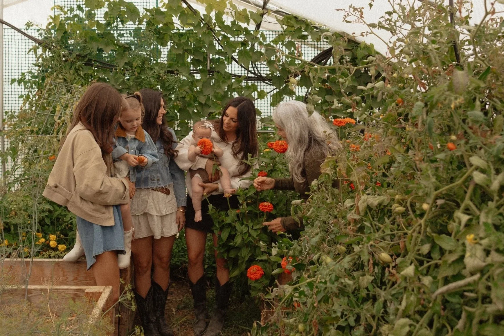 A group of six women, including a baby, in a greenhouse with lush green plants and orange flowers, engaging in flower picking.