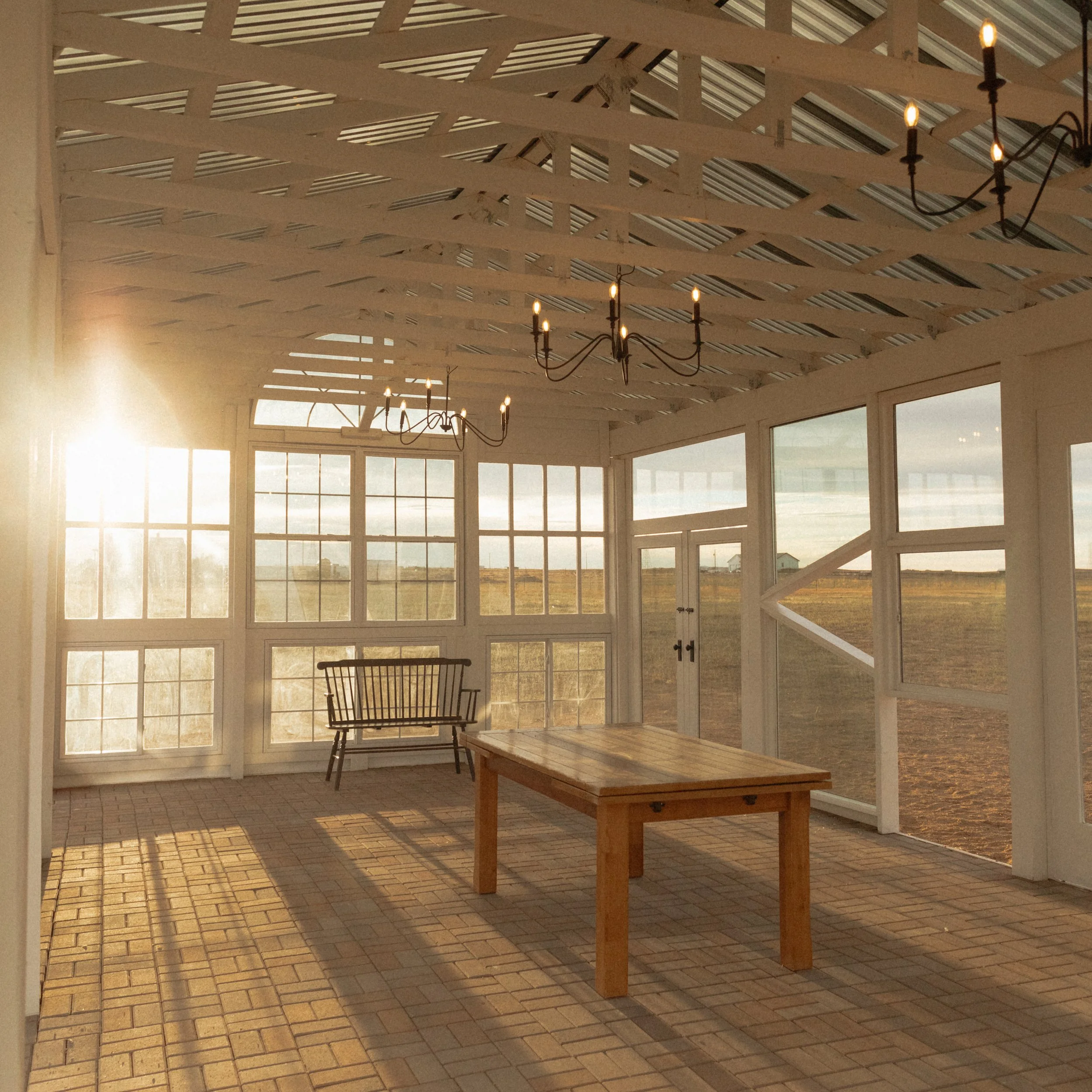 A sunlit indoor space with a brick floor, a wooden table, a wooden chair, large glass windows, a wrought iron chandelier with candles, and white painted wooden beams on the ceiling.