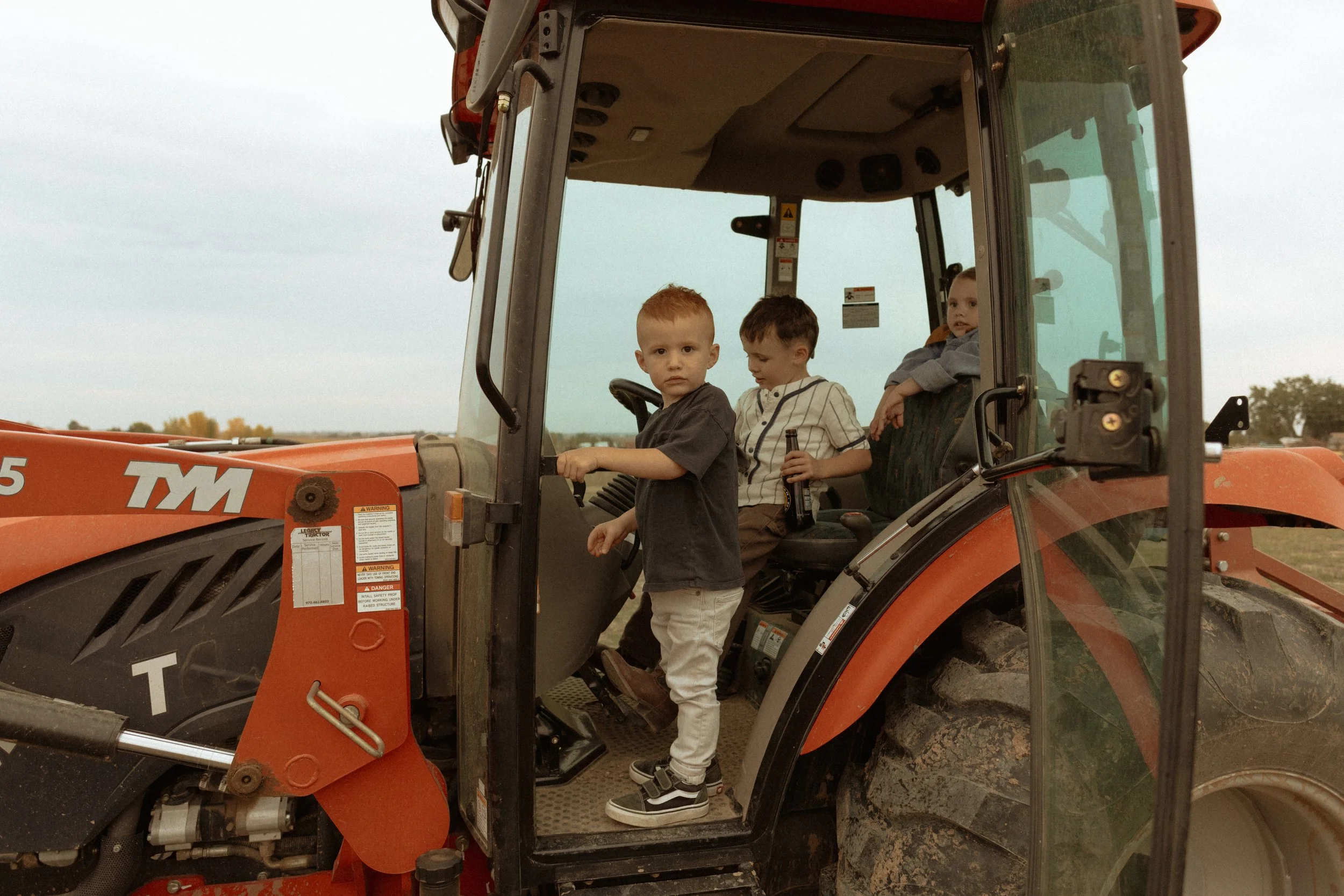 Three children sitting on a tractor with one child standing in the driver’s seat, outdoors in a field.