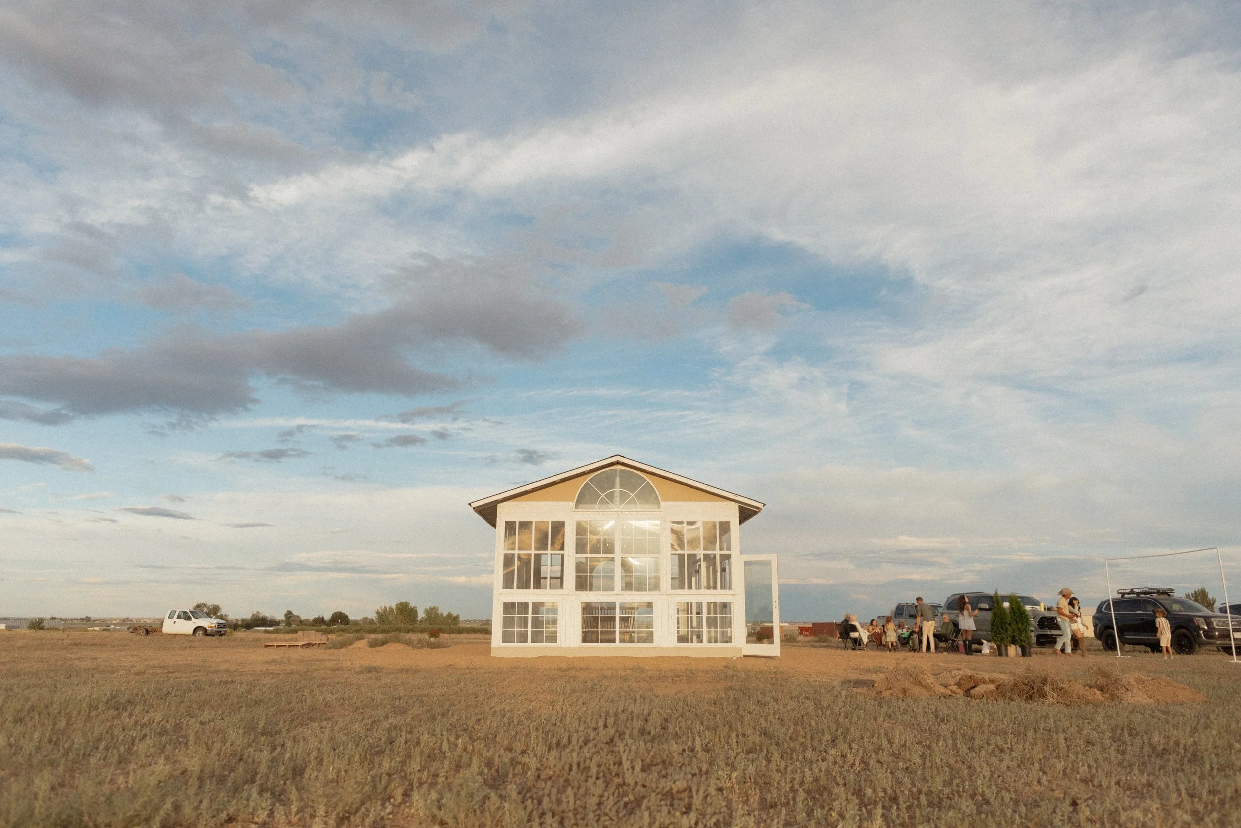 A glass greenhouse with a wooden frame stands in an open field under a partly cloudy sky, with a group of people and parked cars nearby.