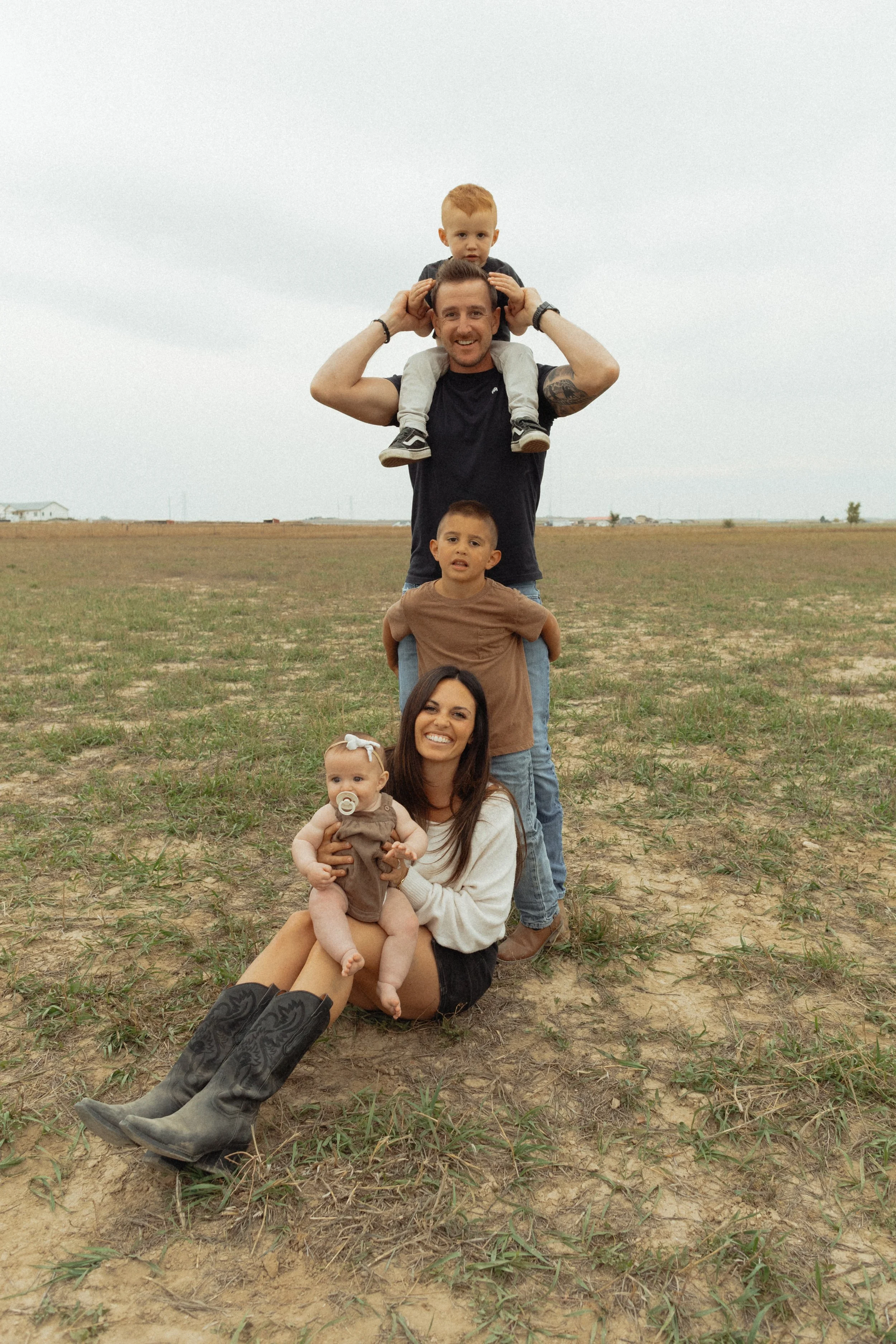 A family of five poses outdoors on a cloudy day with an open field behind them. The man, with tattoos, is standing and smiling while carrying a young boy on his shoulders. A woman is sitting on the ground holding a baby girl in her lap. The family appears happy and relaxed.