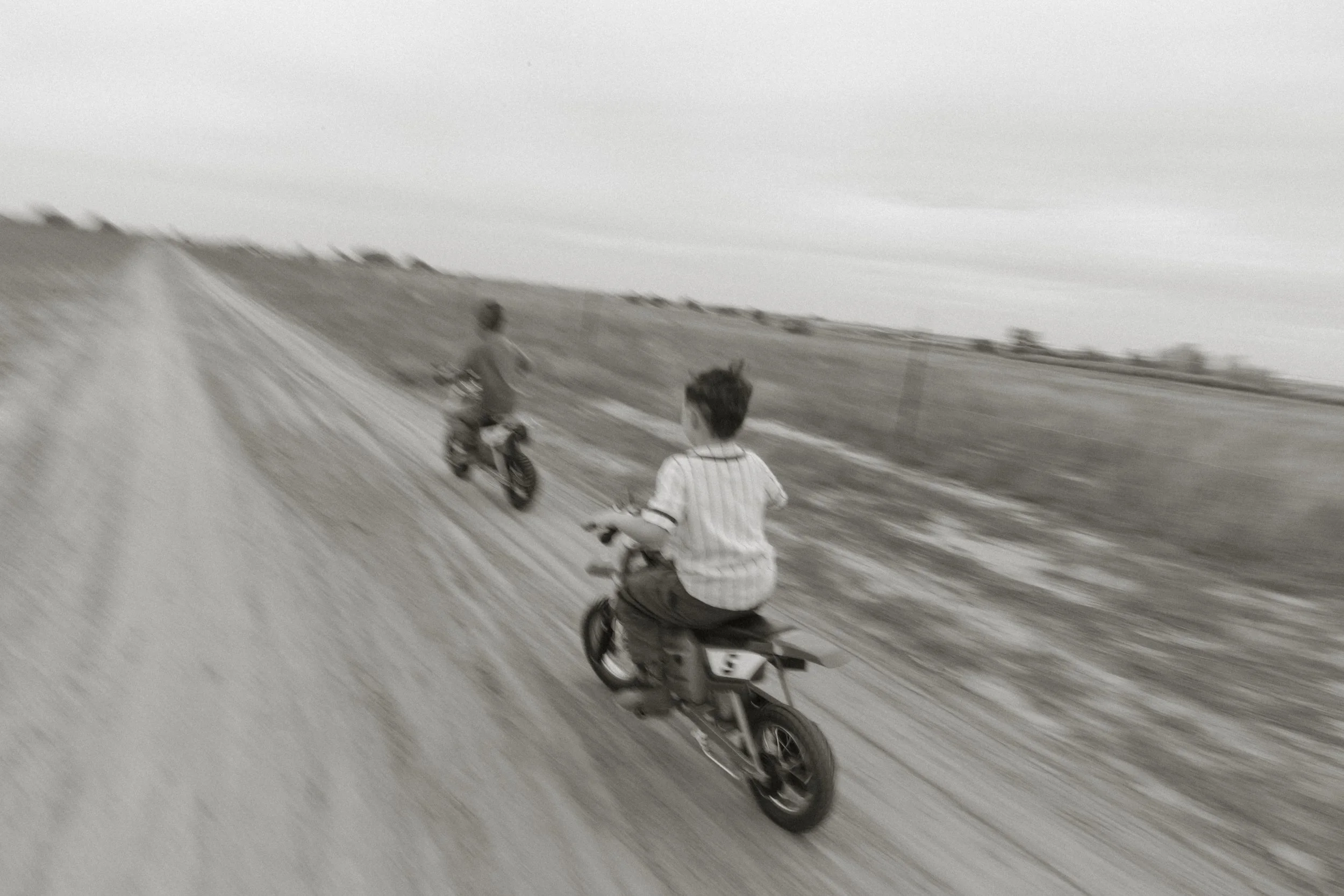 Two children riding bicycles on a dirt path in a rural area, with grassy fields and rolling hills in the background.