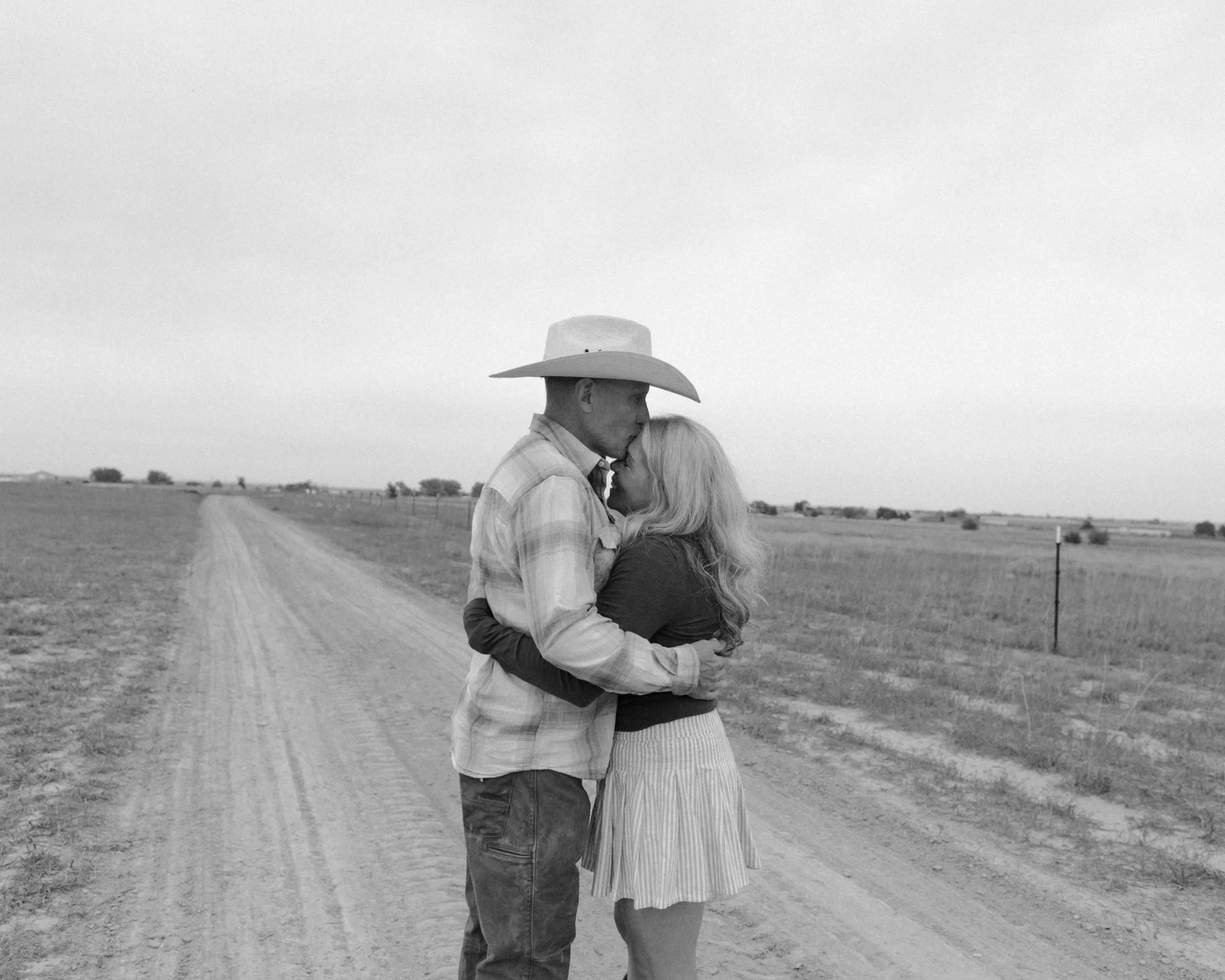A black and white photo of a couple kissing in an open rural landscape, with the man wearing a cowboy hat and the woman with long hair, standing on a dirt road.