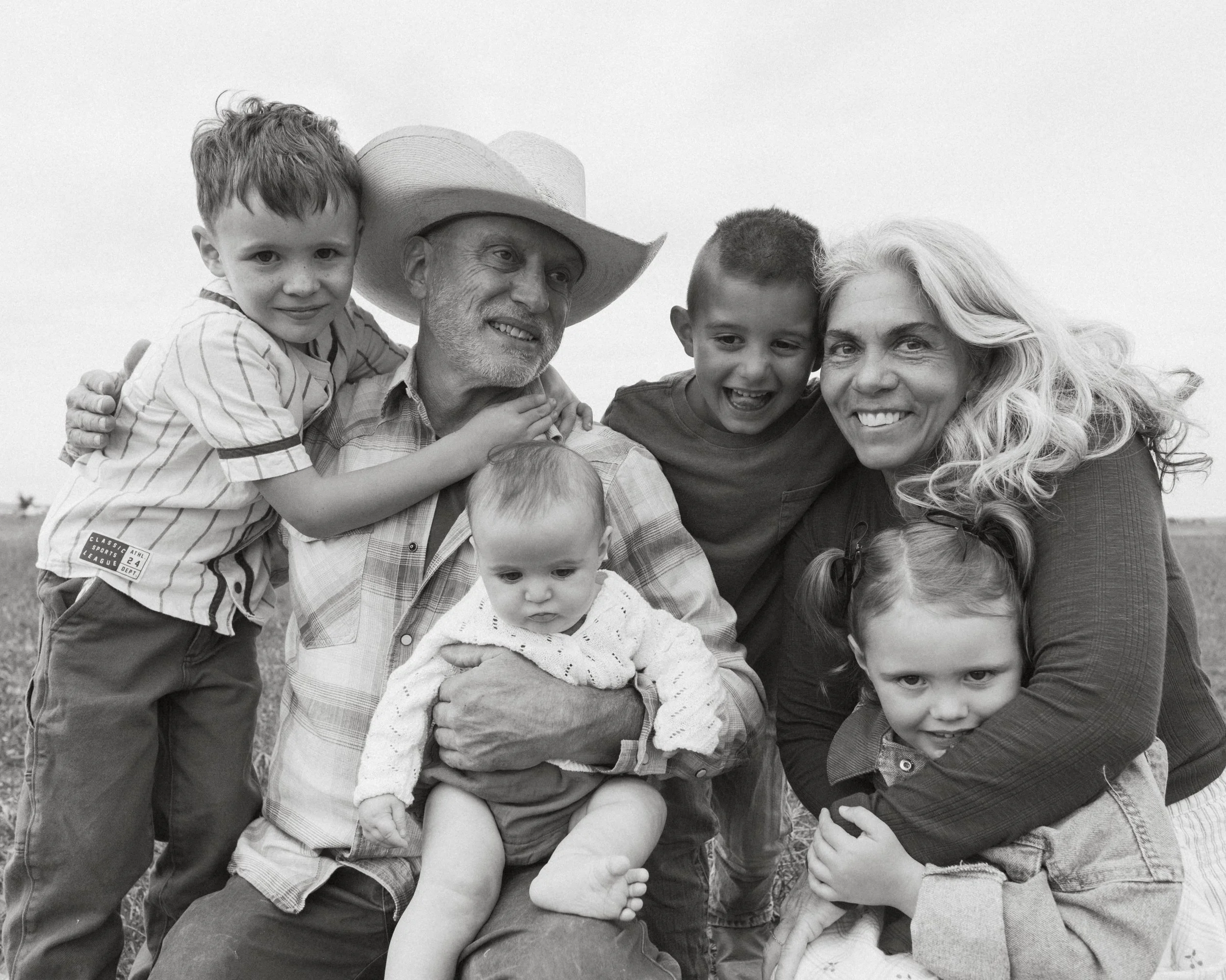 A group of a woman, an older man wearing a cowboy hat, and five children smiles for a photo outdoors. The woman has long, wavy hair and the children are of various ages, some sitting and some being held.