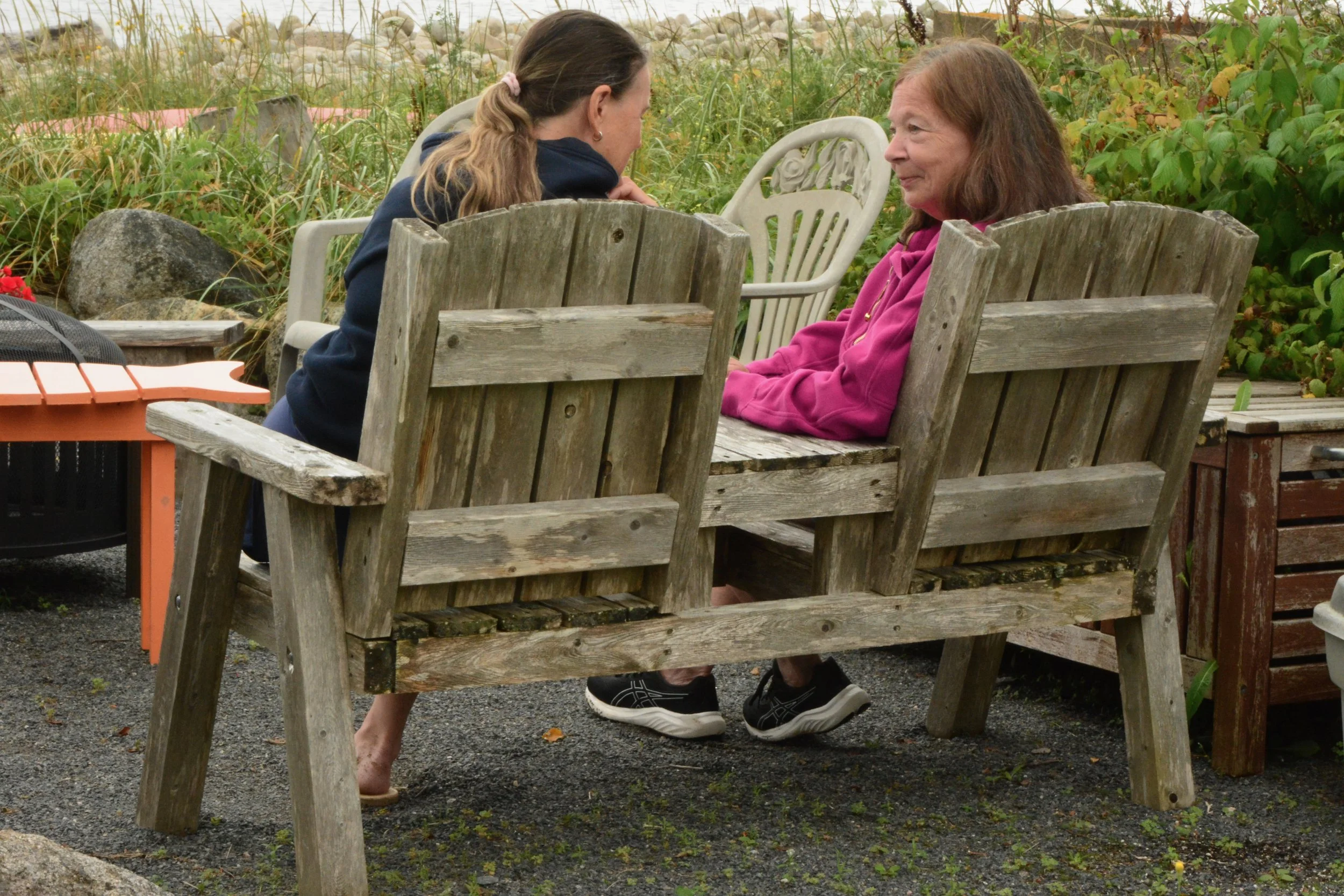 Two women sitting on wooden benches outdoors, leaning toward each other, engaged in conversation. One woman has long brown hair and is wearing a black hoodie, while the other has shoulder-length brown hair and is wearing a pink jacket. There are plants and rocks in the background.