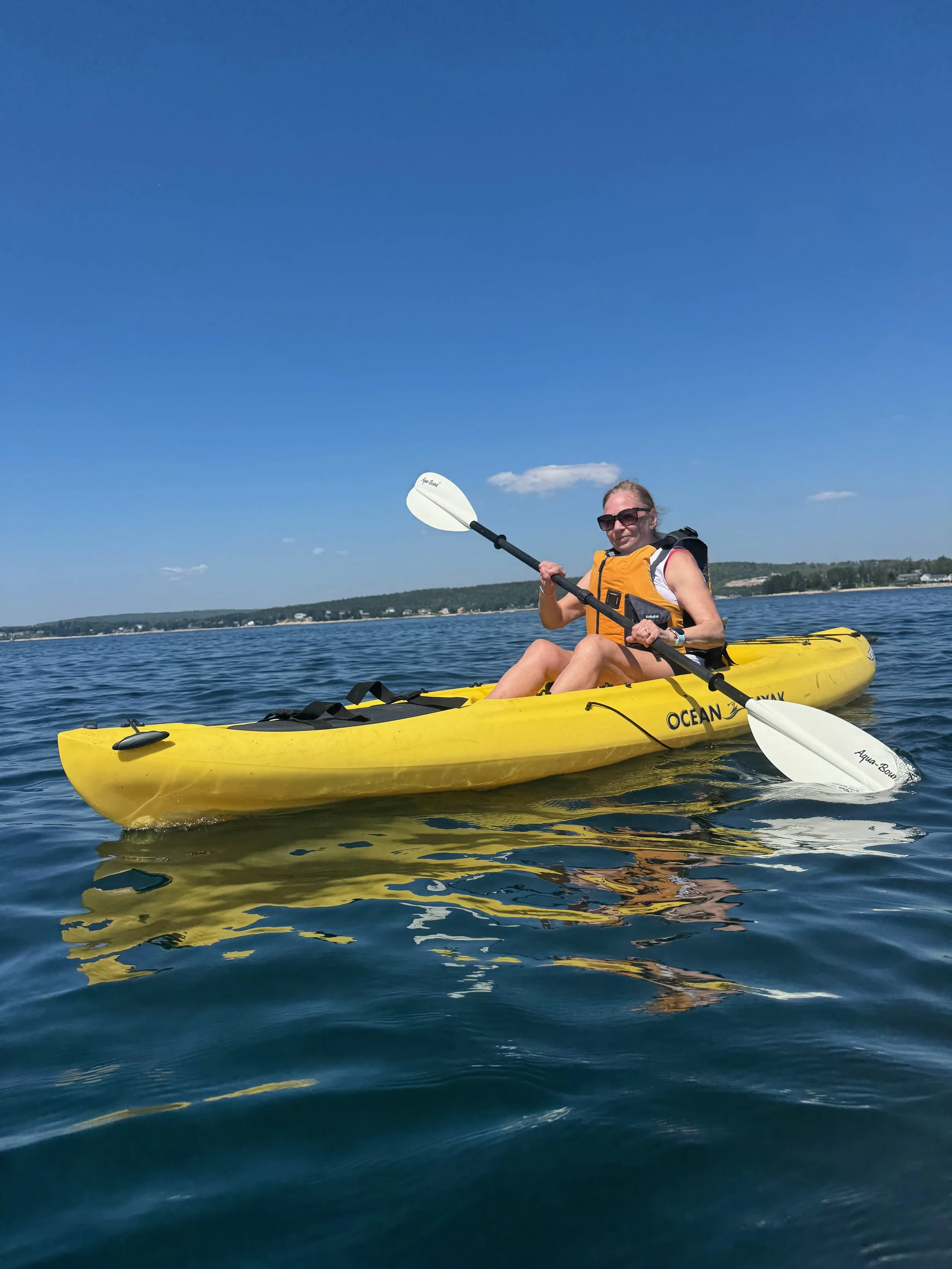 Woman wearing sunglasses and a yellow life jacket kayaking on a calm body of water with distant shoreline and blue sky