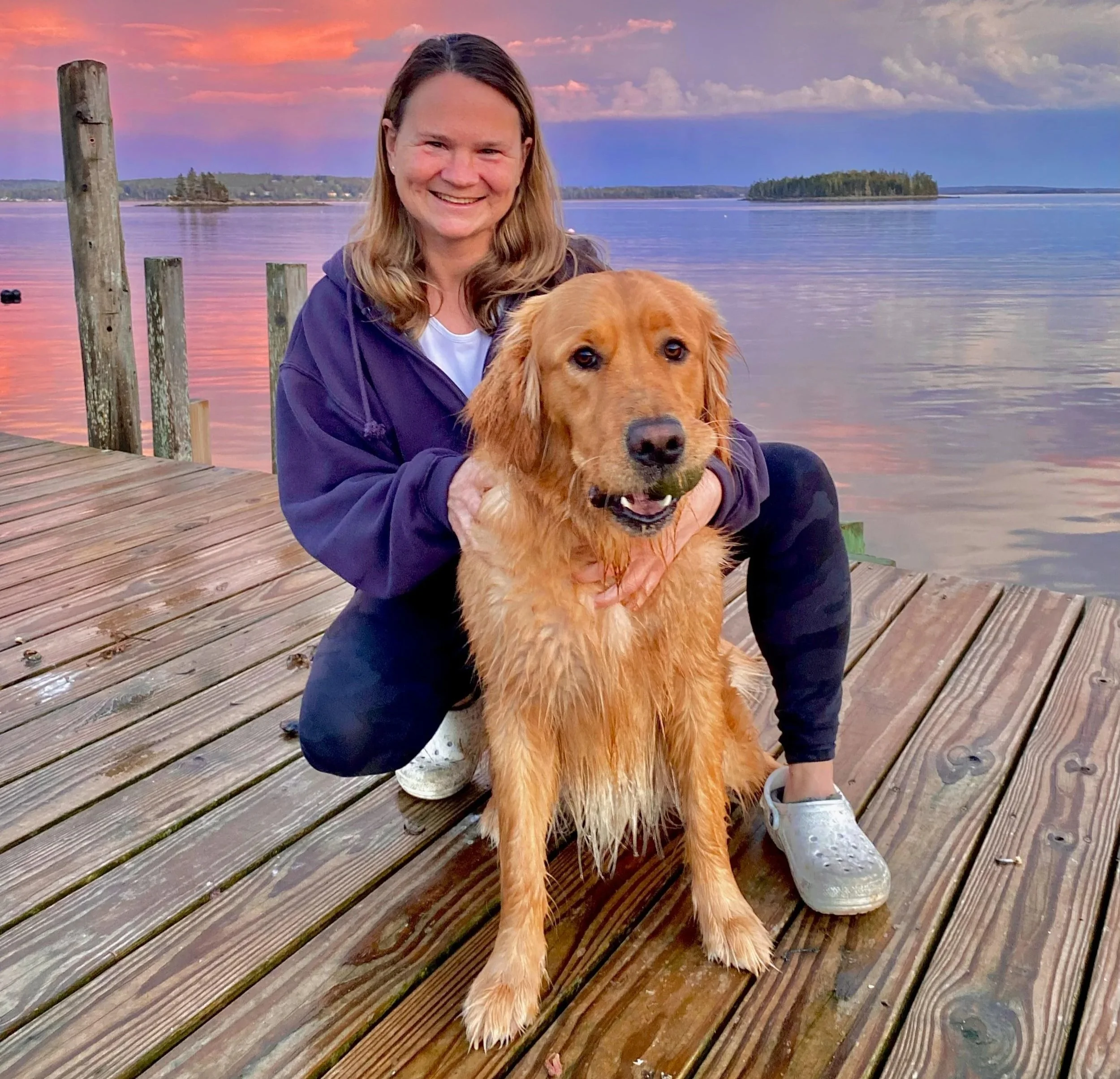 A smiling woman with shoulder-length hair wearing a purple jacket, holding a wet golden retriever dog on a wooden dock over a lake during sunset, with two small islands and a colorful sky with pink and purple clouds in the background.