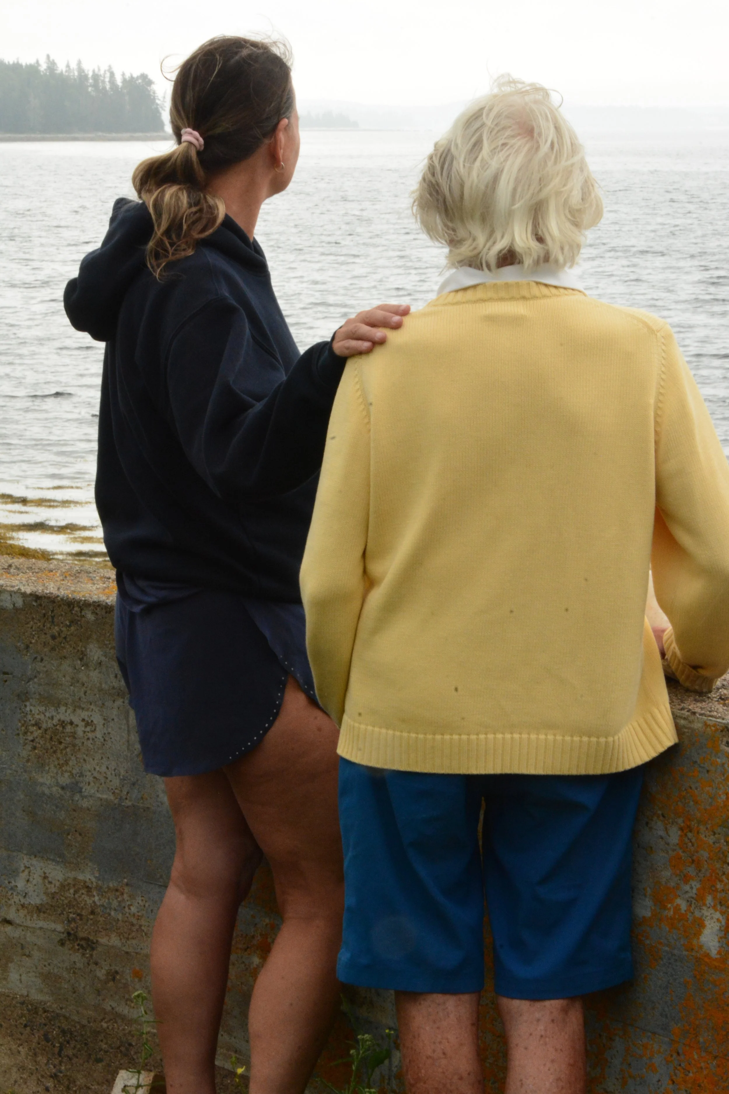 Two women stand near a body of water, facing away from the camera, with one woman resting her hand on the other's shoulder.