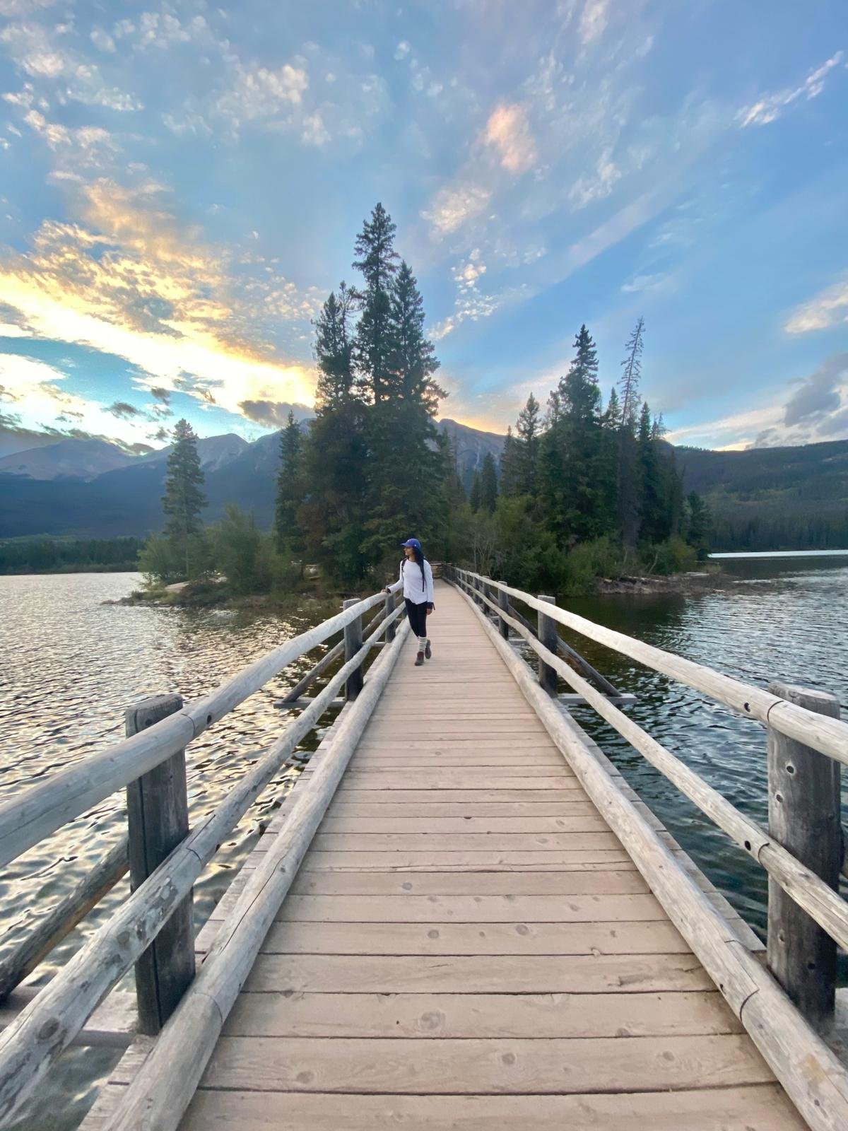 A woman in a white jacket, dark pants, and a blue hat walking on a wooden bridge over a lake, surrounded by trees, with mountains and a partly cloudy sky in the background at sunset.