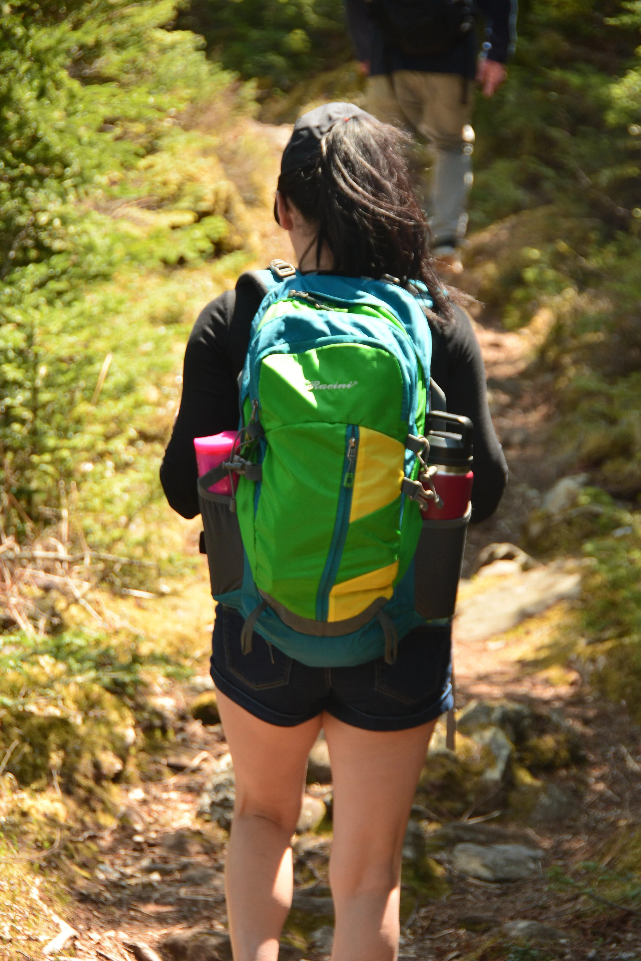 A woman hiking on a trail in a forest, wearing a green and blue backpack, black shorts, and a black top, with another person walking ahead.