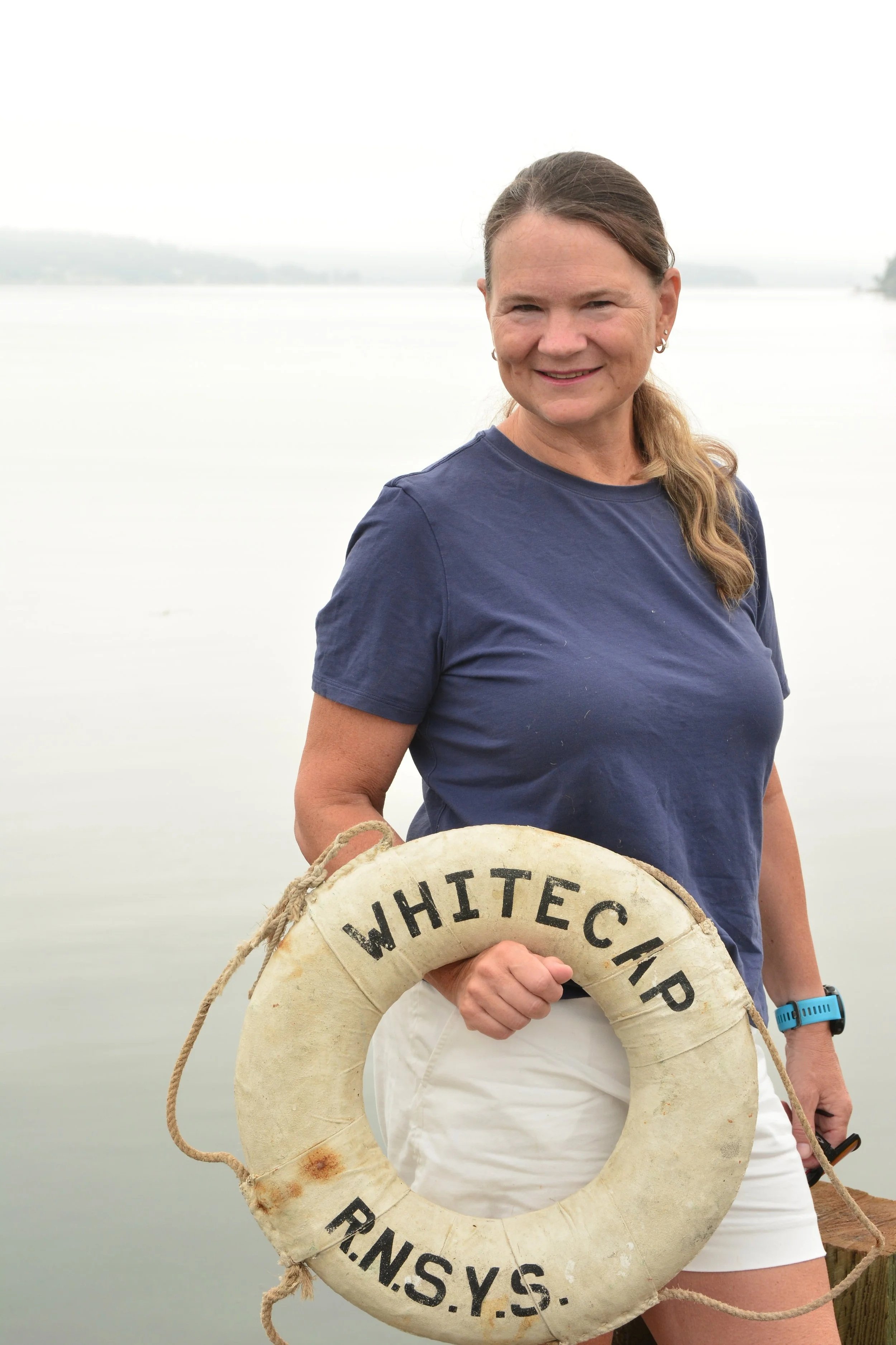 A woman with long hair tied back, wearing a navy blue shirt, smiling while holding a white lifebuoy with black lettering that reads 'WHITECAP R.N.S.Y.S.' She is standing near a body of water with a foggy or overcast sky in the background.