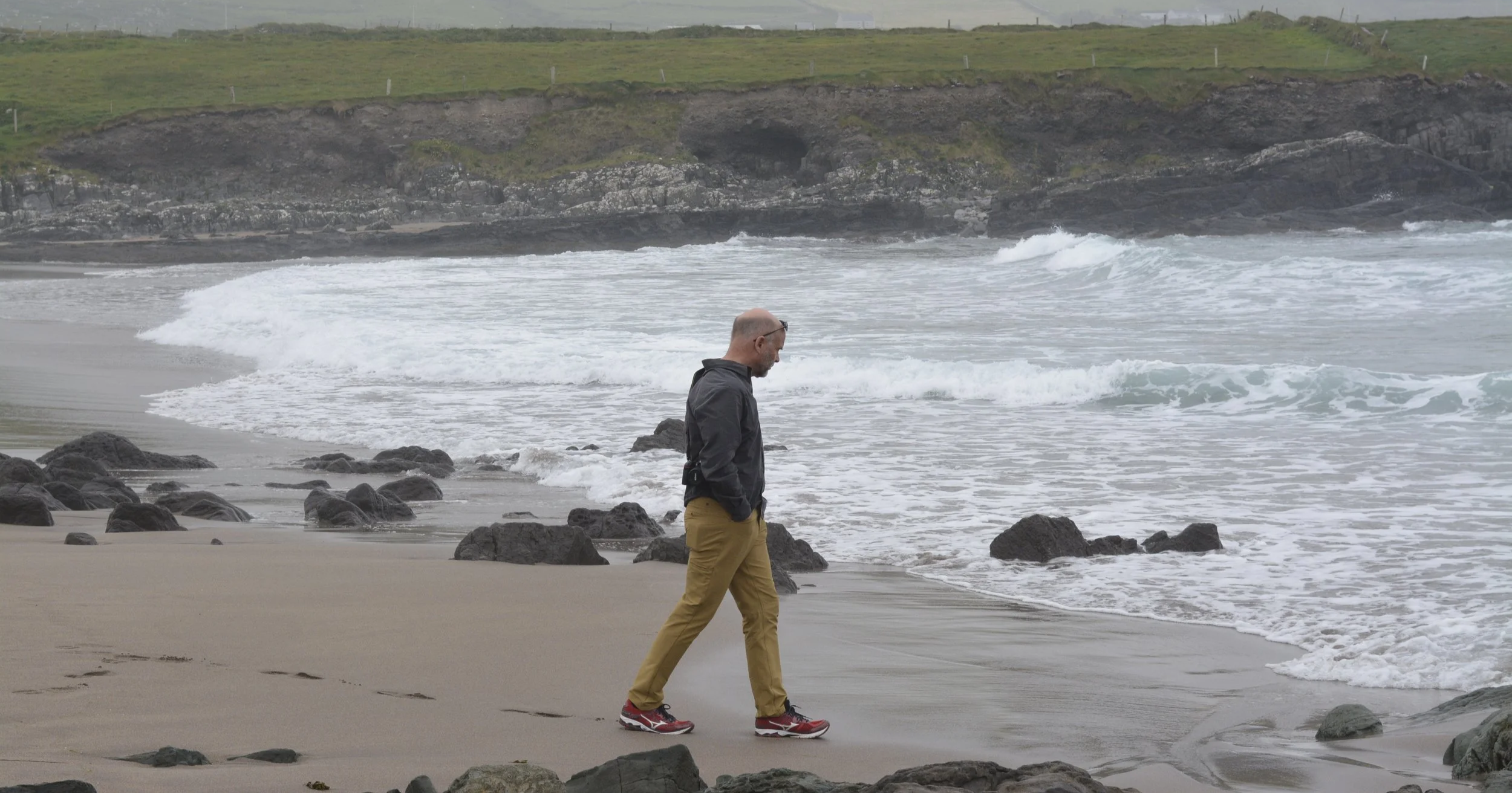 A man walking along the sandy shore of a beach with rocks, looking down at the ocean waves, with grassy cliffs in the background under an overcast sky.