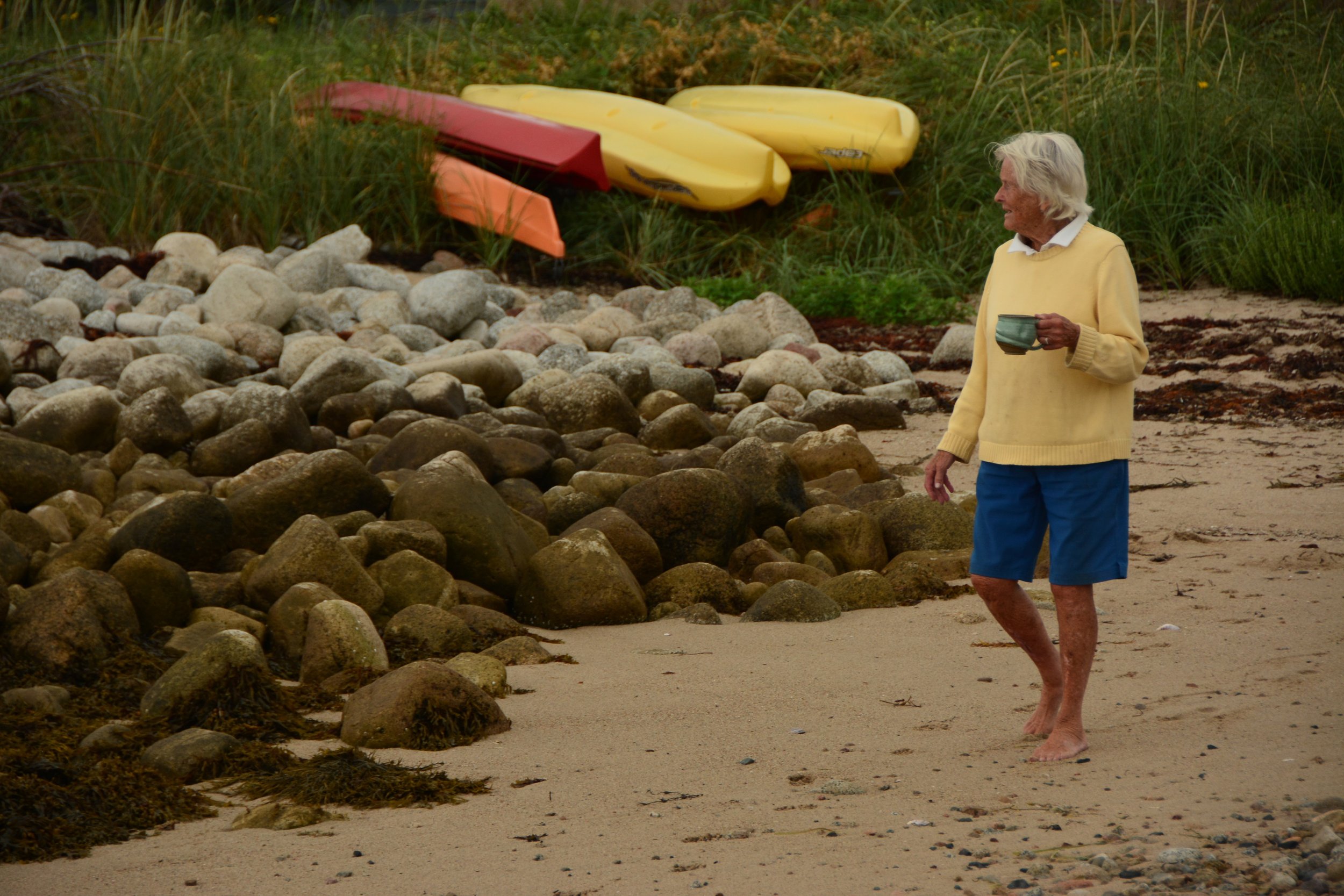 An elderly woman with white hair, wearing a yellow sweater and blue shorts, walking barefoot on a sandy beach holding a mug. Behind her, there are colorful kayaks on the grass next to large rocks.