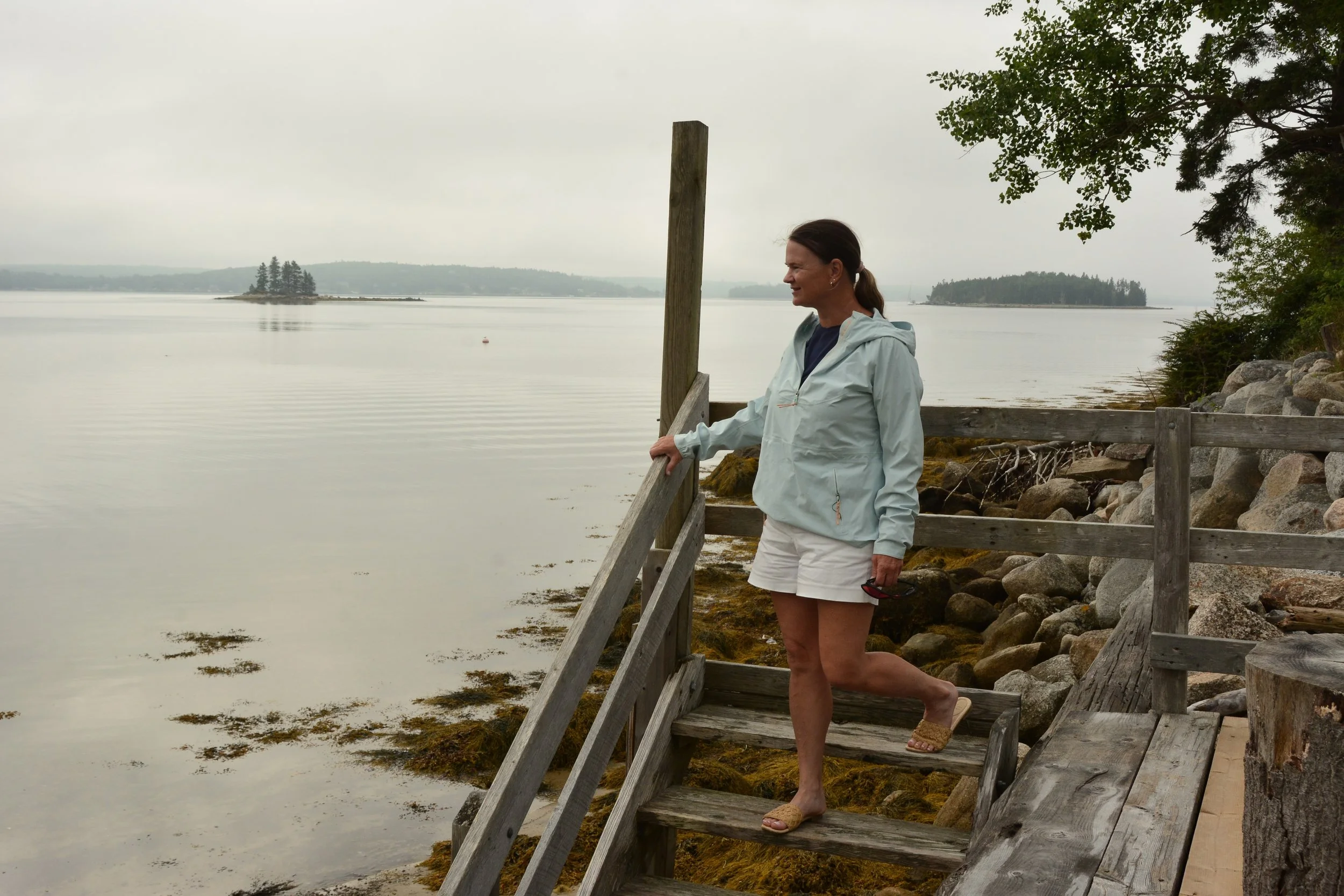 A woman standing on a wooden dock by the water, with trees and islands in the background, overcast sky.