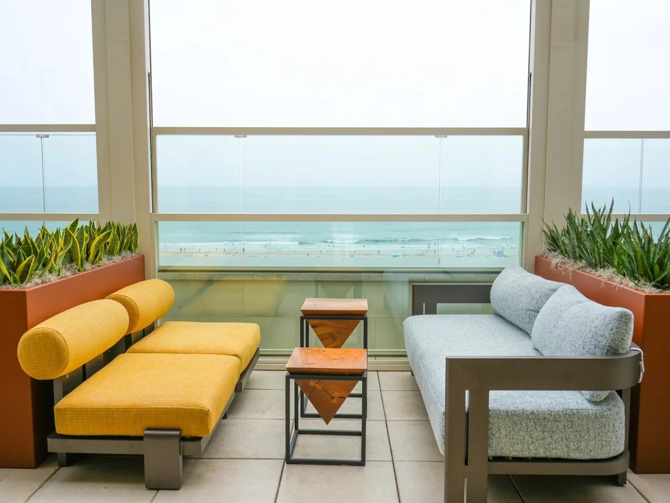 Oceanfront lounge seating with yellow and blue chairs, framed by glass walls and modern planters overlooking the beach.