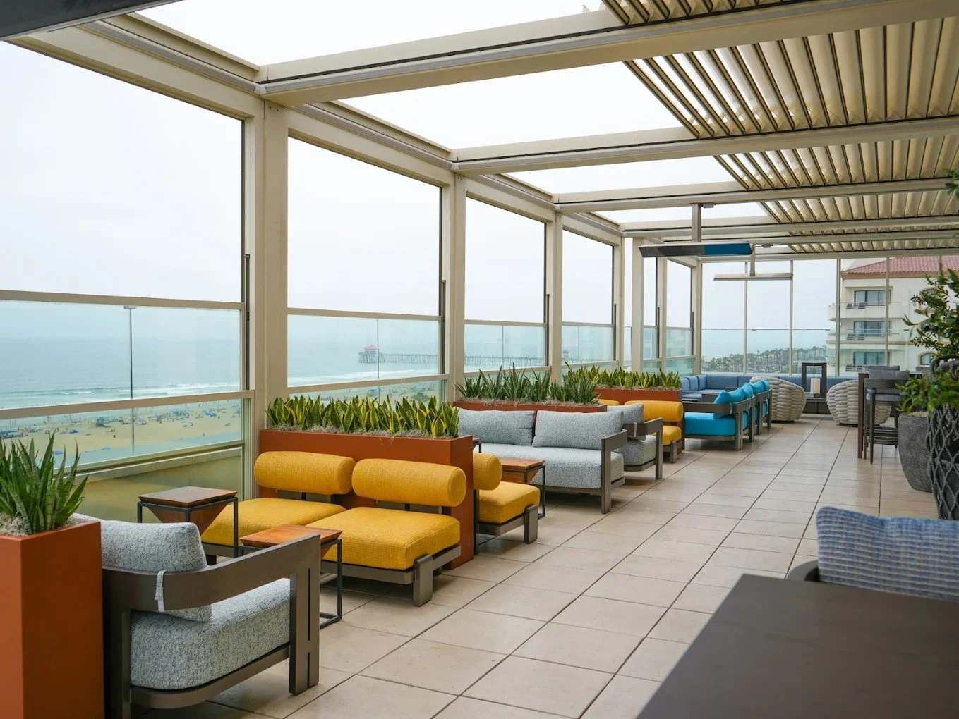 Expansive rooftop lounge showing multiple seating zones, planters, and adaptive louvered roofing with a panoramic ocean backdrop.