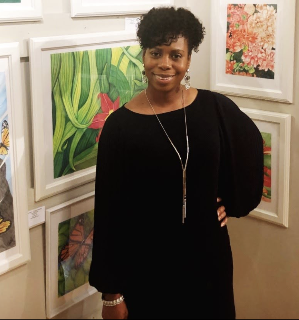 A woman with short, curly black hair wearing a black dress with long sleeves, earrings, a necklace, and a bracelet standing in front of colorful artwork at an art gallery.