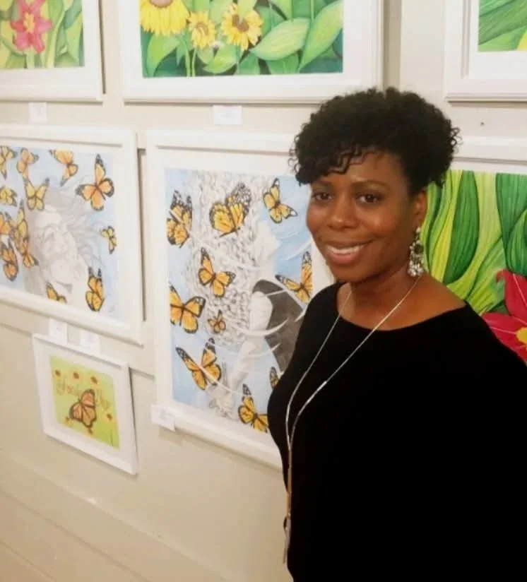 A woman smiling at an art gallery, standing in front of framed paintings featuring butterflies, flowers, and other colorful nature scenes.
