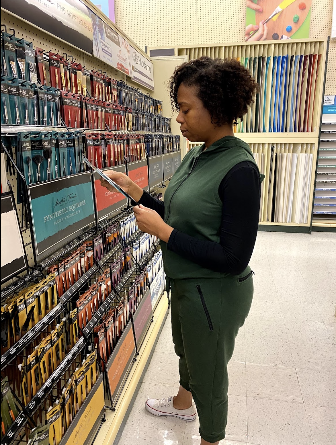 A woman shopping for paint brushes in a store aisle, standing next to organized paintbrushes and paint supplies.