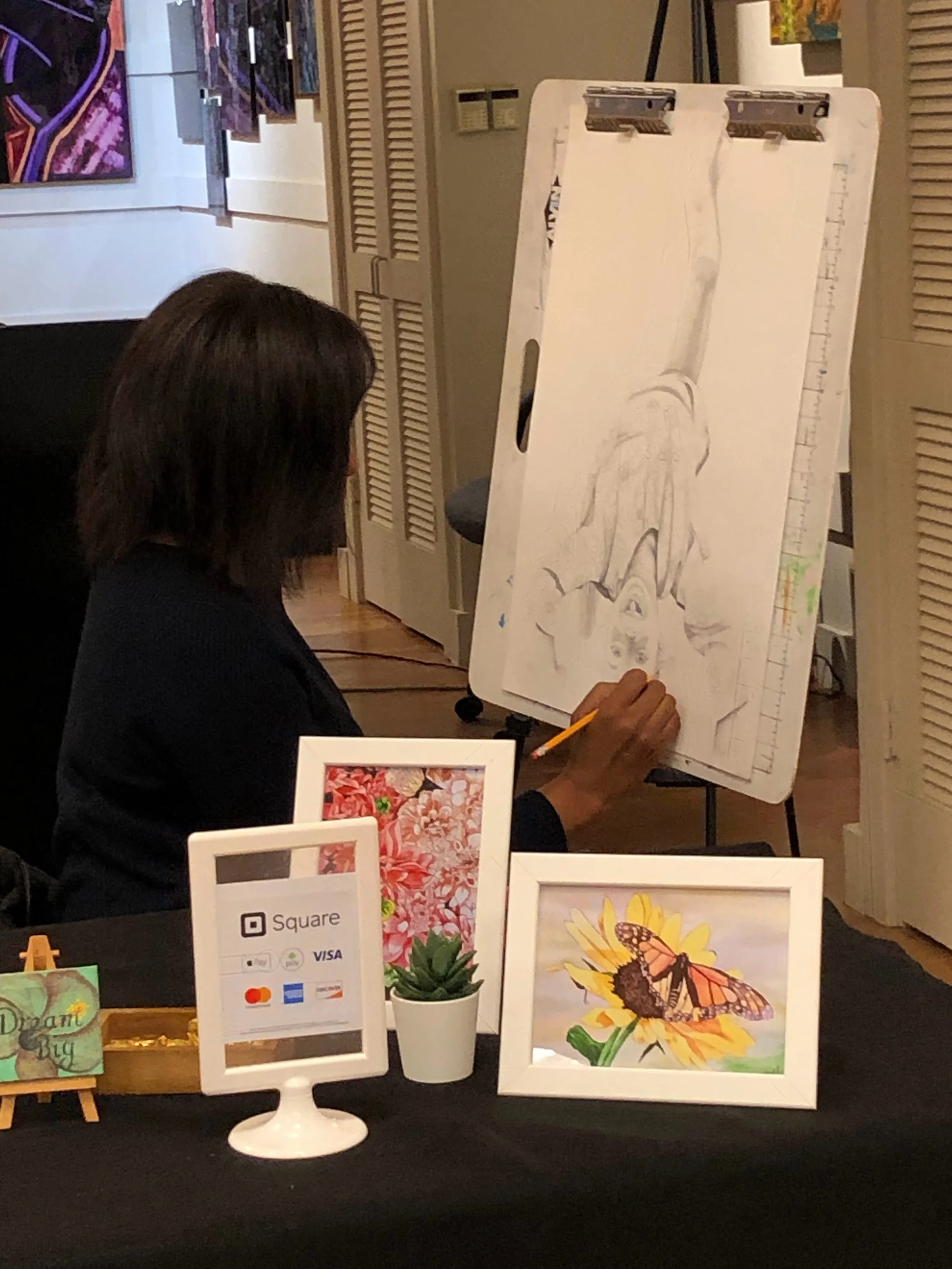 An art display featuring watercolor illustrations of a butterfly on a sunflower, a sunflower, and a pink flower, along with a small cropping of a geometric design on another artwork in the background. There is a card with payment logos and a small potted plant on the table.