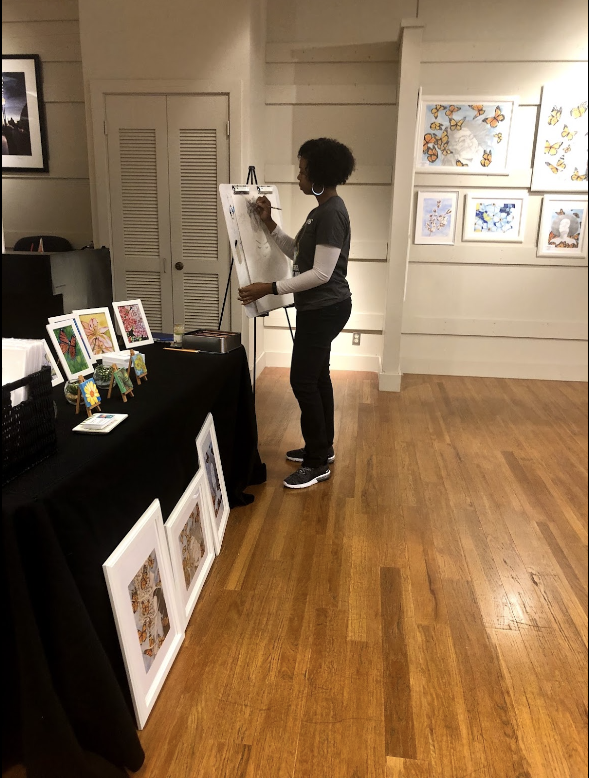 A woman with curly hair drawing on a whiteboard at an art display in a gallery. Art pieces, including butterfly images, are on the wall and table.