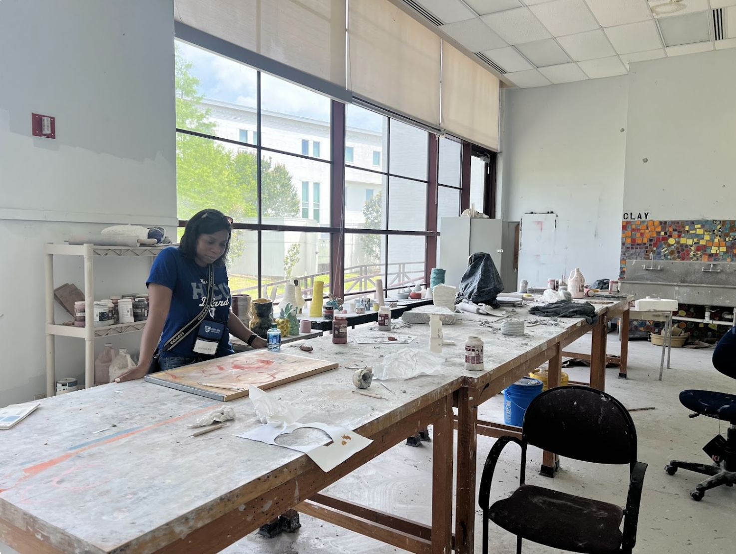 An art classroom with a large wooden table covered in art supplies and pottery. A woman is standing at the table looking down, with large windows behind her showing trees and a building outside. The room appears to be used for ceramics or pottery, with painted tiles on the wall and a sink in the background.