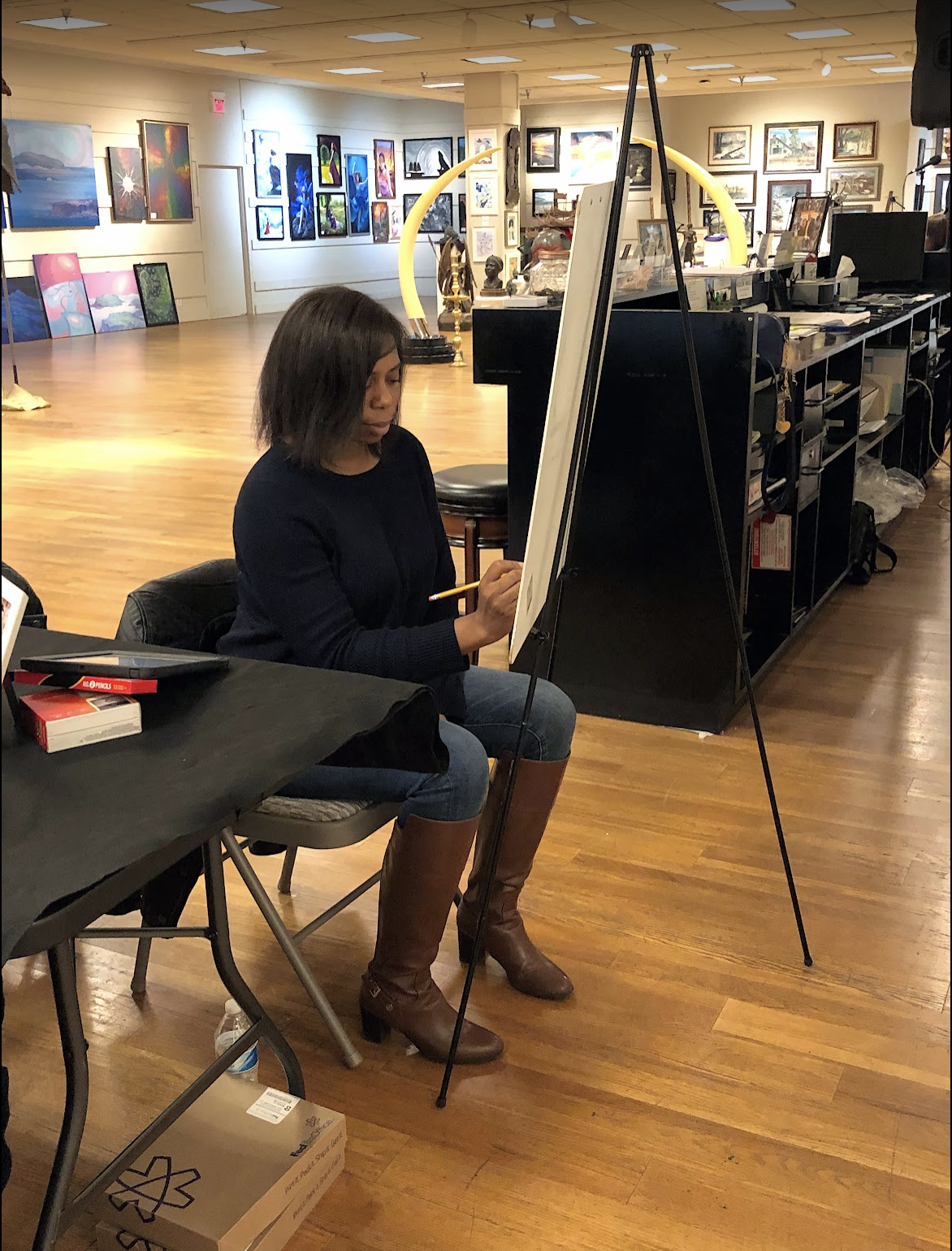 A woman painting on a canvas at an art gallery or studio, with many framed artworks displayed on the walls behind her.