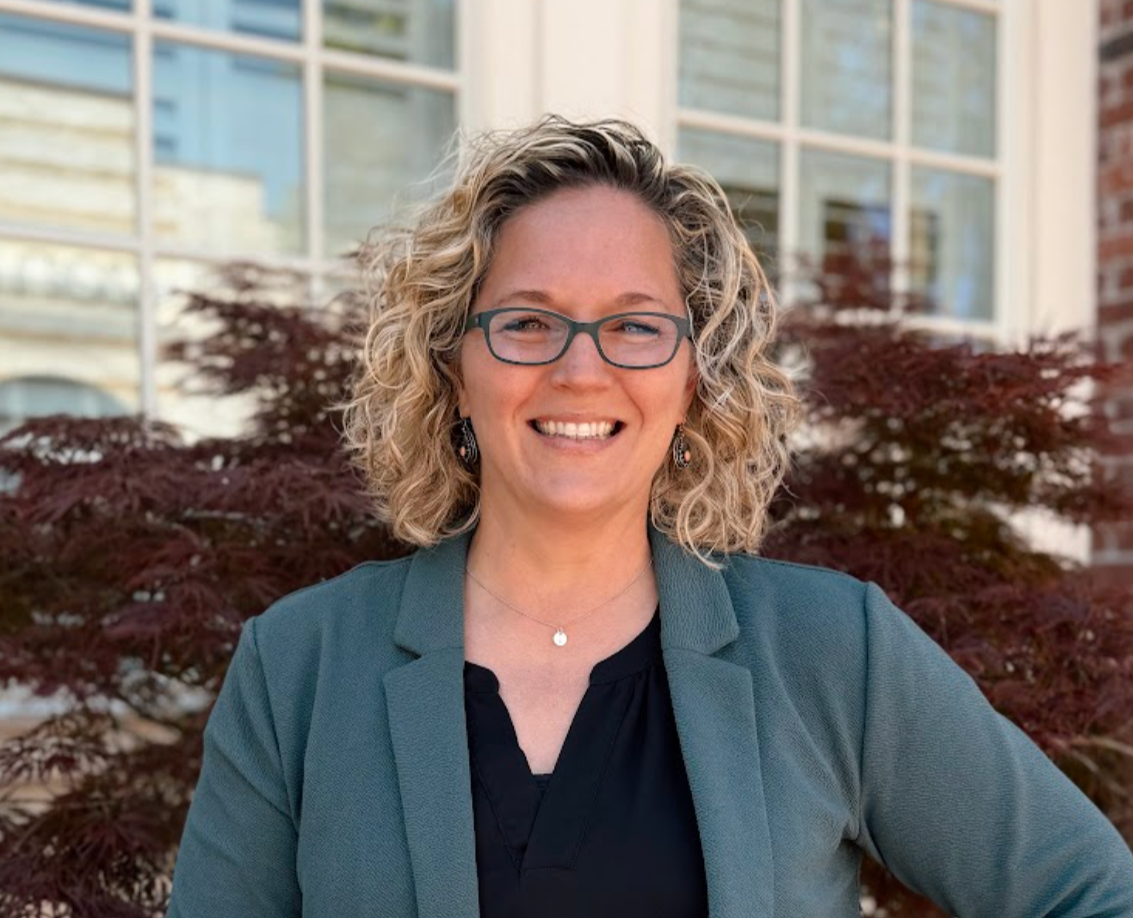 A woman with curly blonde hair and glasses, smiling, standing outdoors in front of a building with large windows and red-leafed bushes.