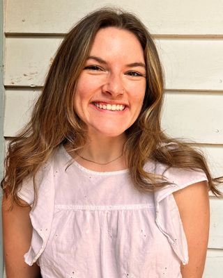 A woman with long brown hair smiling in front of a white wooden wall.