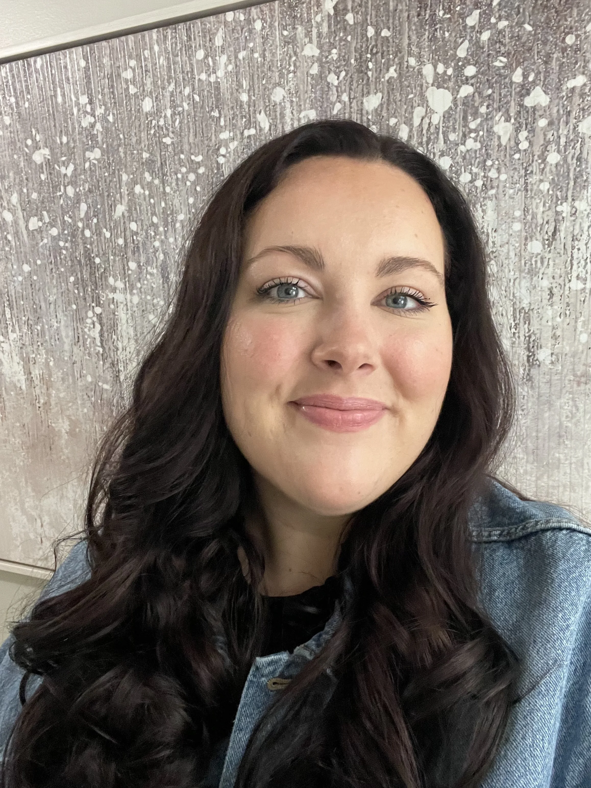 Close-up portrait of a woman with long, wavy dark brown hair, blue eyes, and light skin, smiling softly in front of a textured silver and white wall.