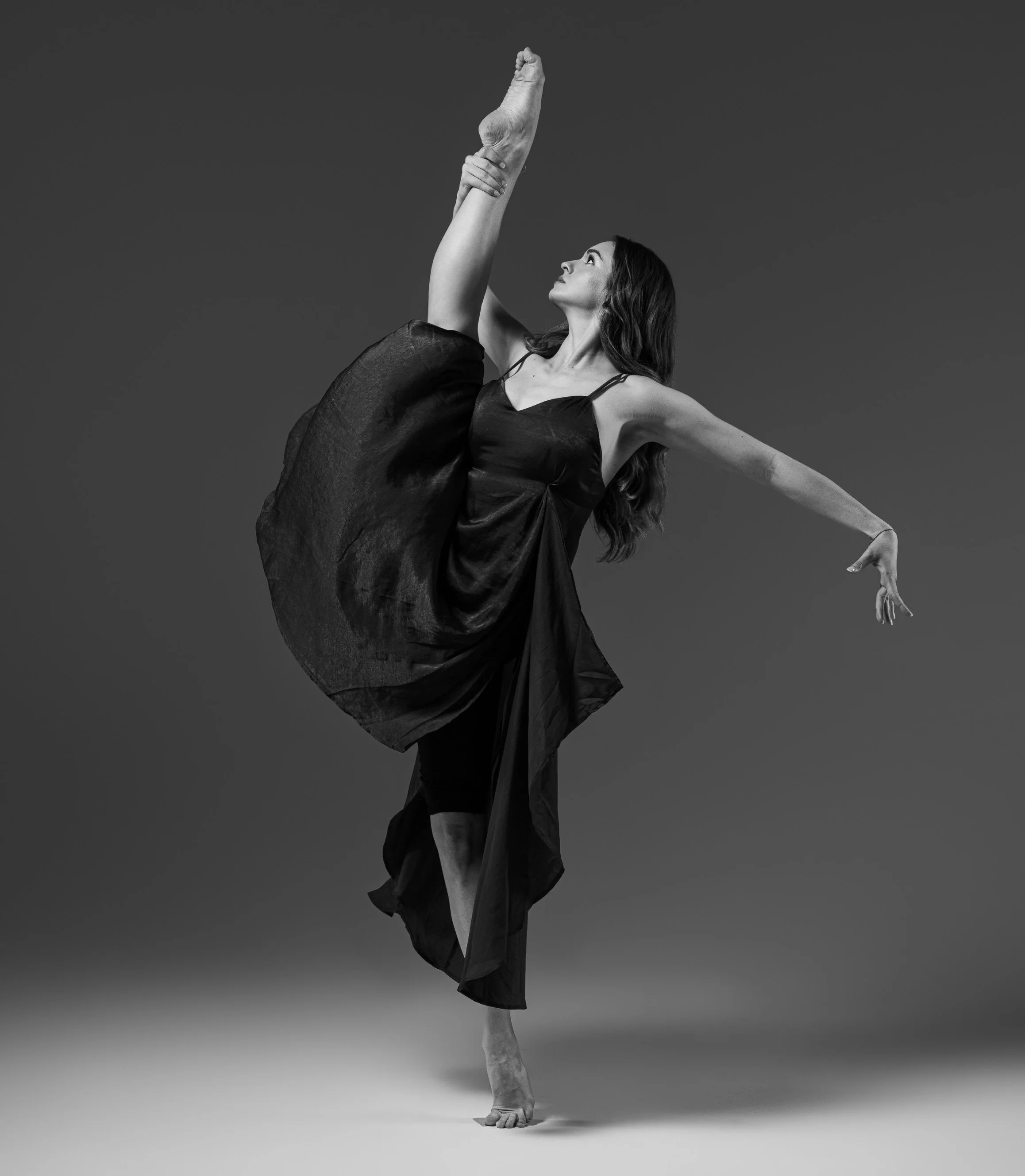 A woman with long hair wearing a black dress performing a yoga pose, balancing on one foot with one arm raised upright and holding her foot with her hand, against a plain gray background.