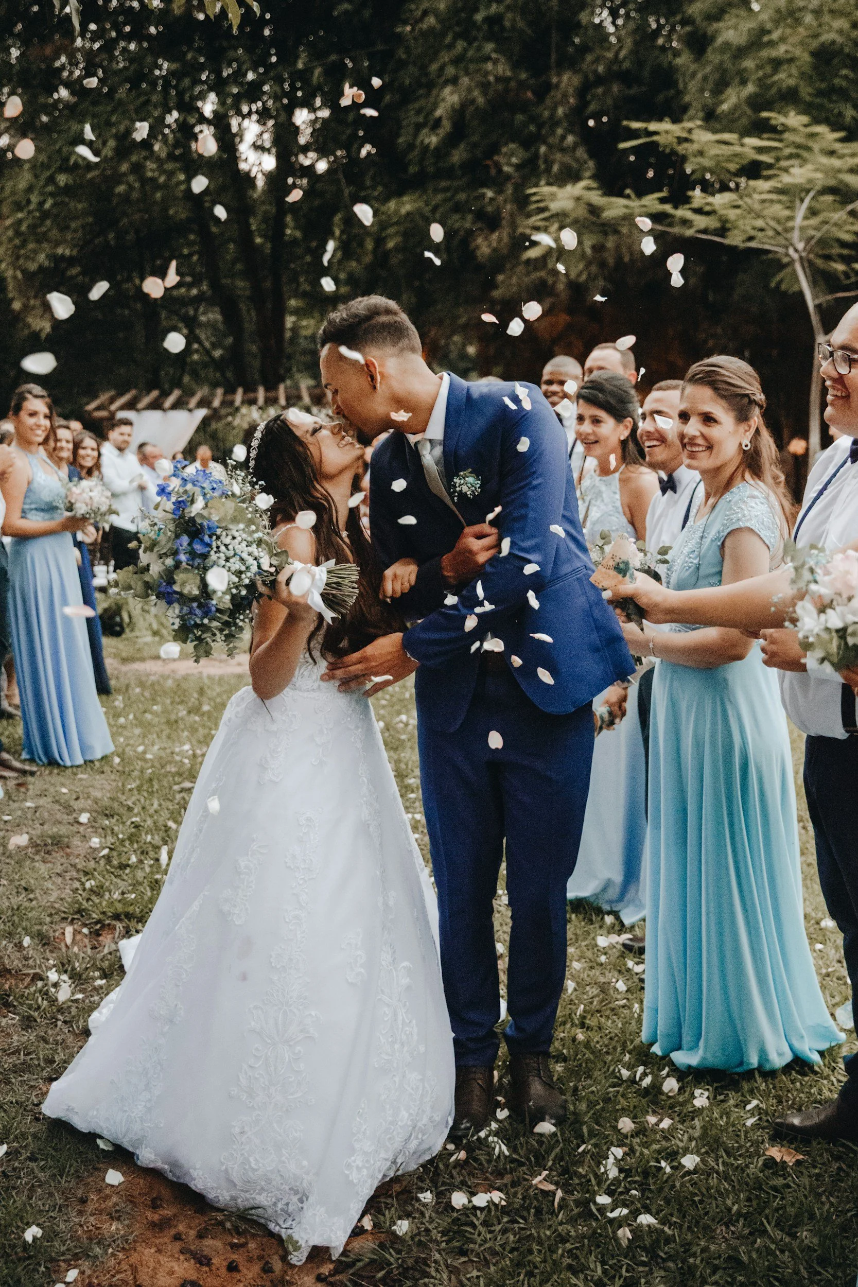 A wedding scene with a bride and groom sharing a kiss while surrounded by friends and family. Flower petals are falling, and some guests are smiling. The bride is holding a bouquet of flowers, and everyone is dressed in formal attire outdoors.