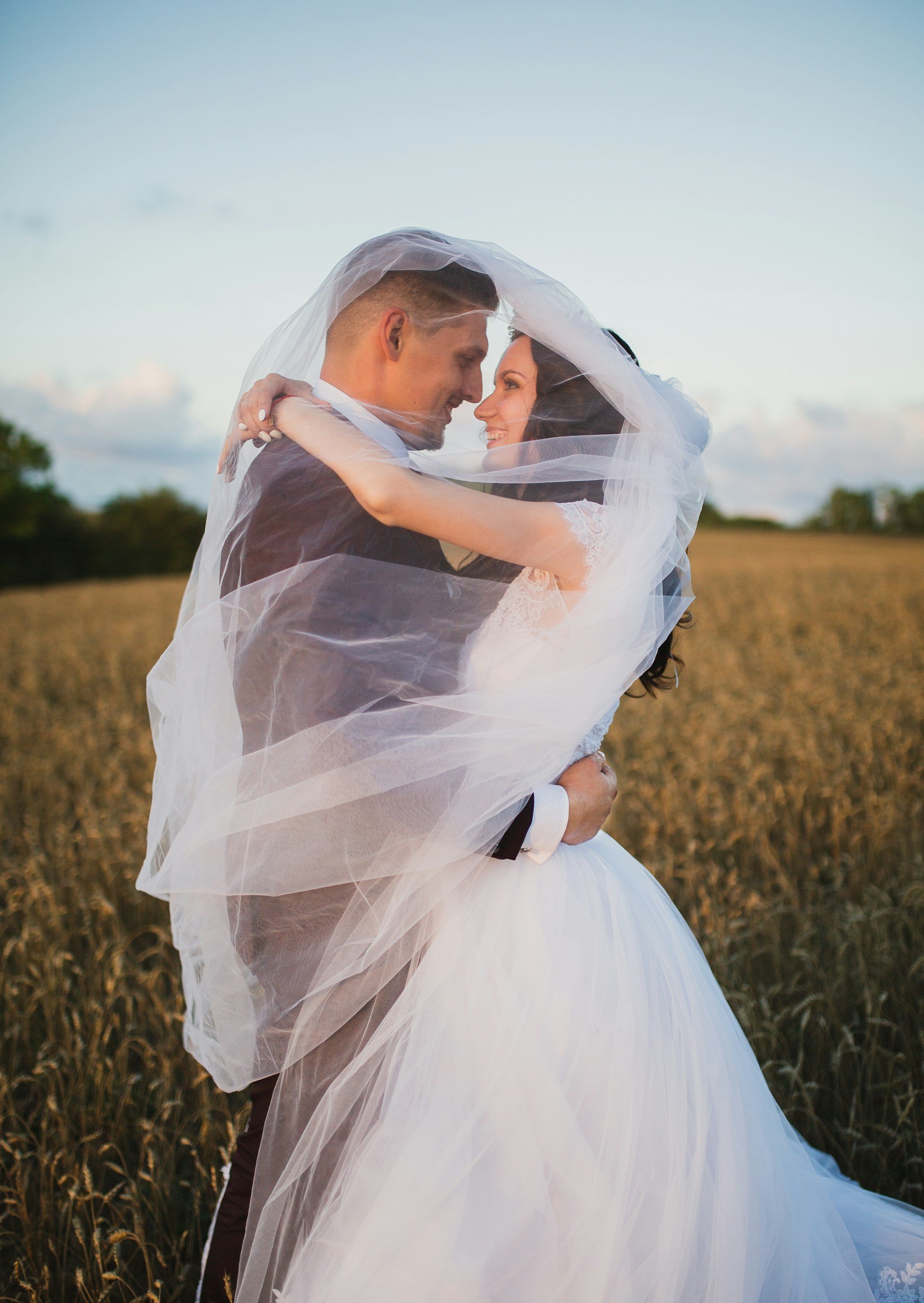 A bride and groom embrace outdoors in a field during sunset, wrapped in a sheer veil.