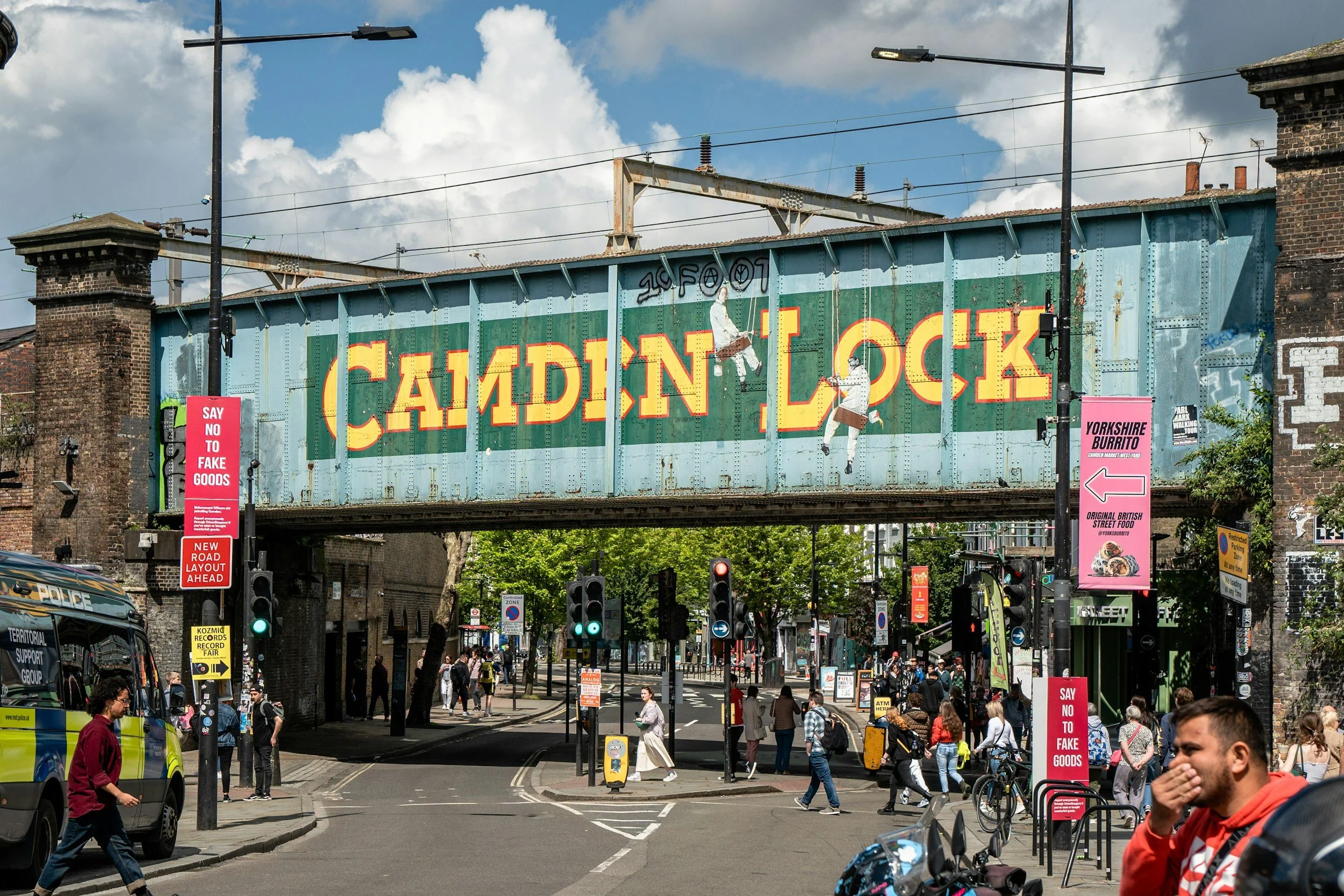 A busy city street scene with pedestrians crossing at a crosswalk, traffic lights, and a bridge with large yellow and red signage reading 'CANDY' and 'LOCK'. There are police vehicles and colorful signs along the street, with historic brick buildings and a cloudy sky overhead.