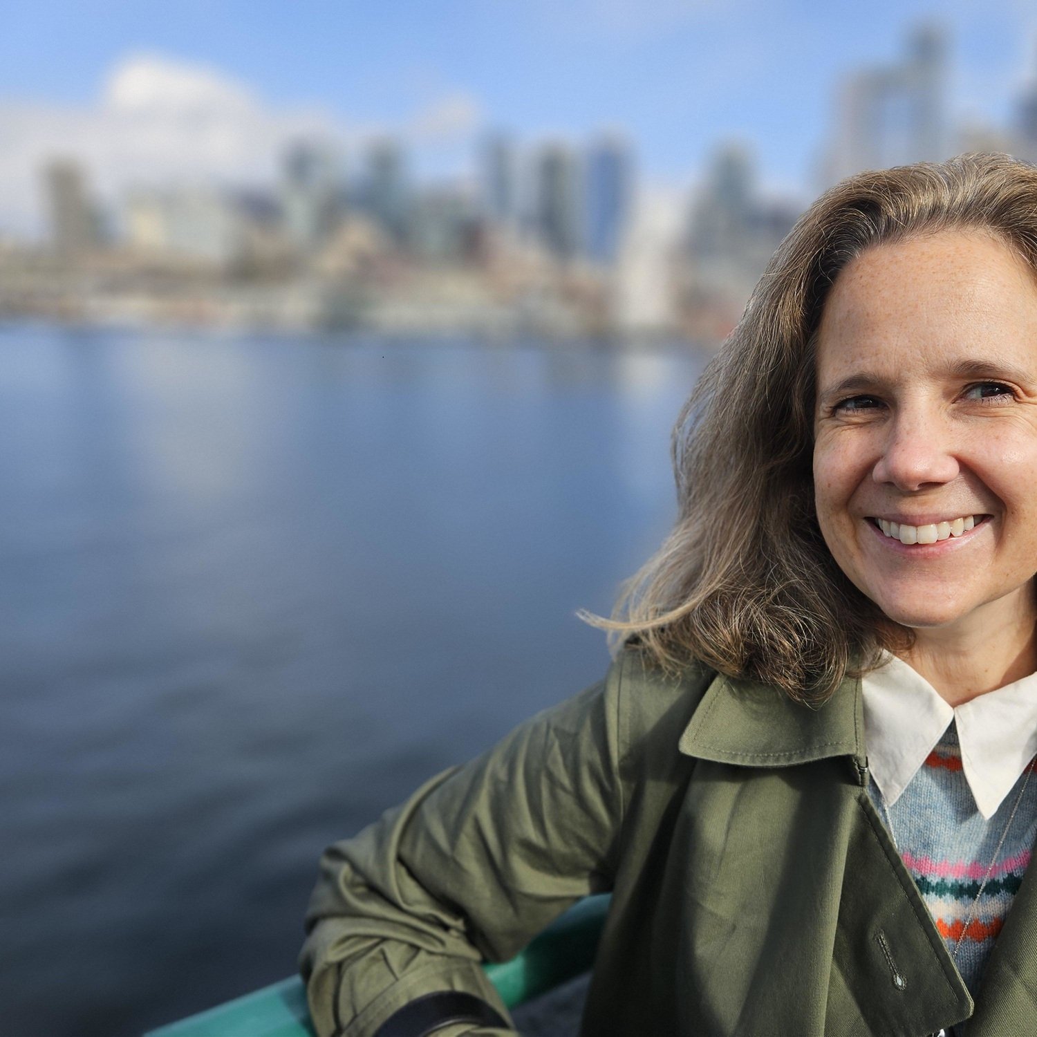A smiling woman with wavy brown hair, wearing a green jacket, sitting outdoors near a body of water with a city skyline in the background.