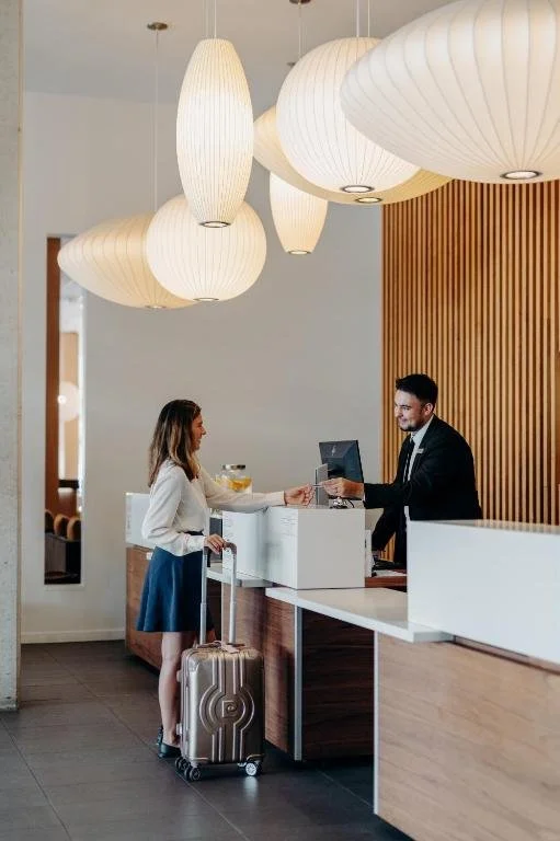 A woman with a rolling suitcase at hotel check-in counter, handing over a card to a male clerk inside a modern hotel lobby with hanging white pendant lights and wooden wall accents.
