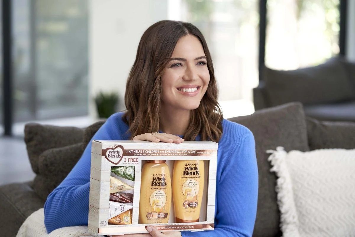 Mandy Moore wearing a blue sweater smiling while holding a gift set of Garnier Whole Blends hair care products in support of UNICEF.
