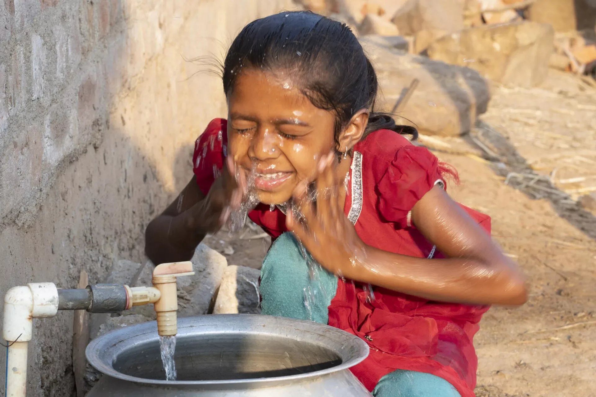 A young girl with wet hair and a red dress is playing at a water tap, splashing water on her face and smiling. The background shows a rural outdoor setting with rocks and dirt.