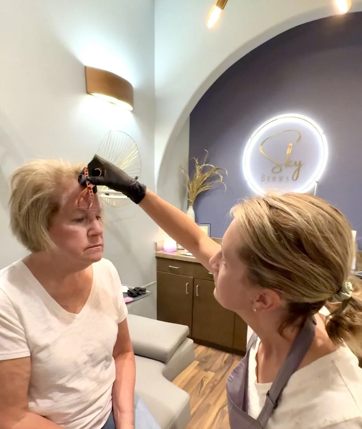 A woman receiving a microblading eyebrow procedure from a technician in a beauty salon, with a sign that reads 'Sky Brows' in the background.