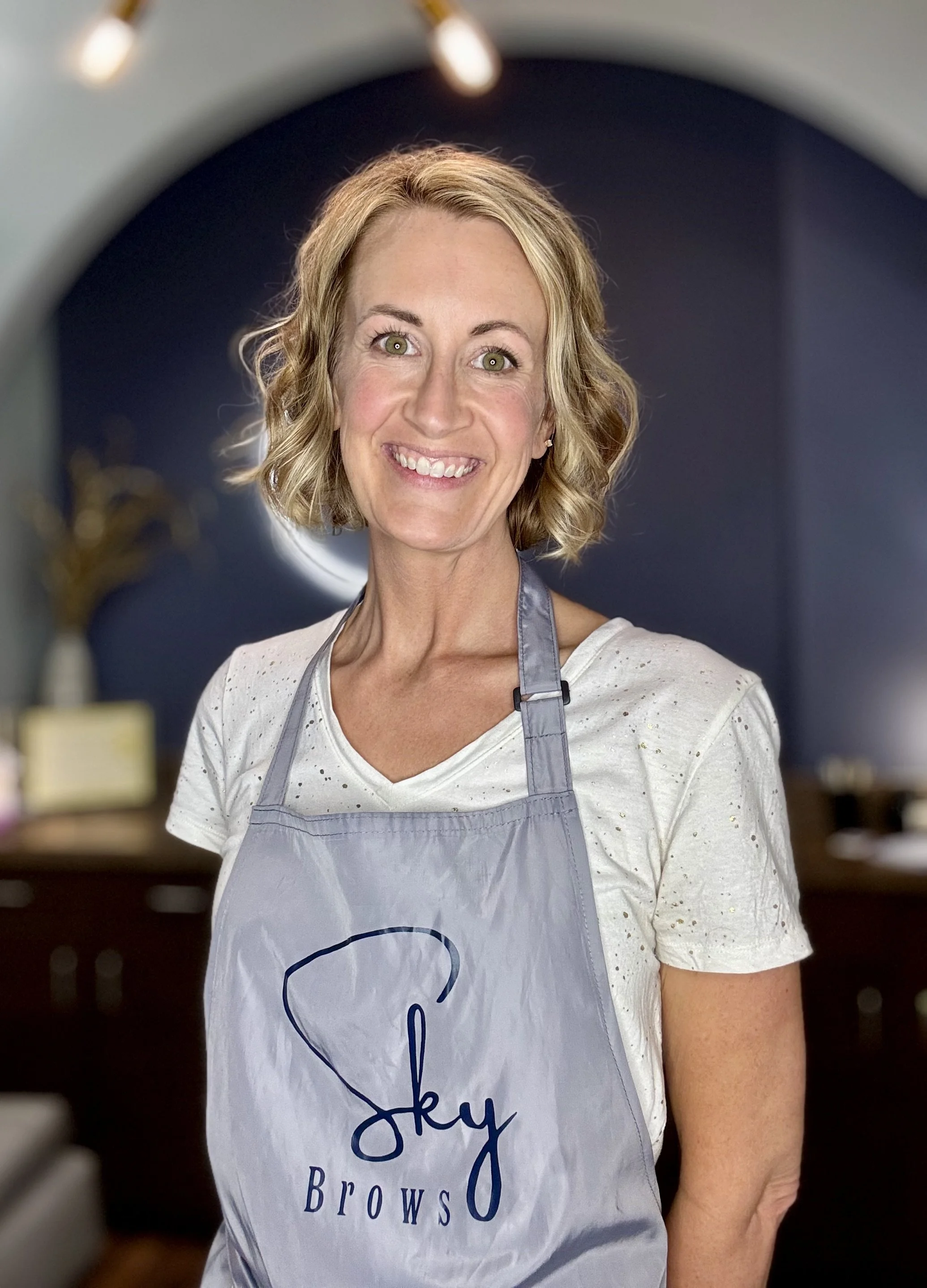 A woman with short blonde hair smiling, wearing a white shirt and a gray apron with a logo that says 'Sky Brows' in blue, standing indoors with blurred background.