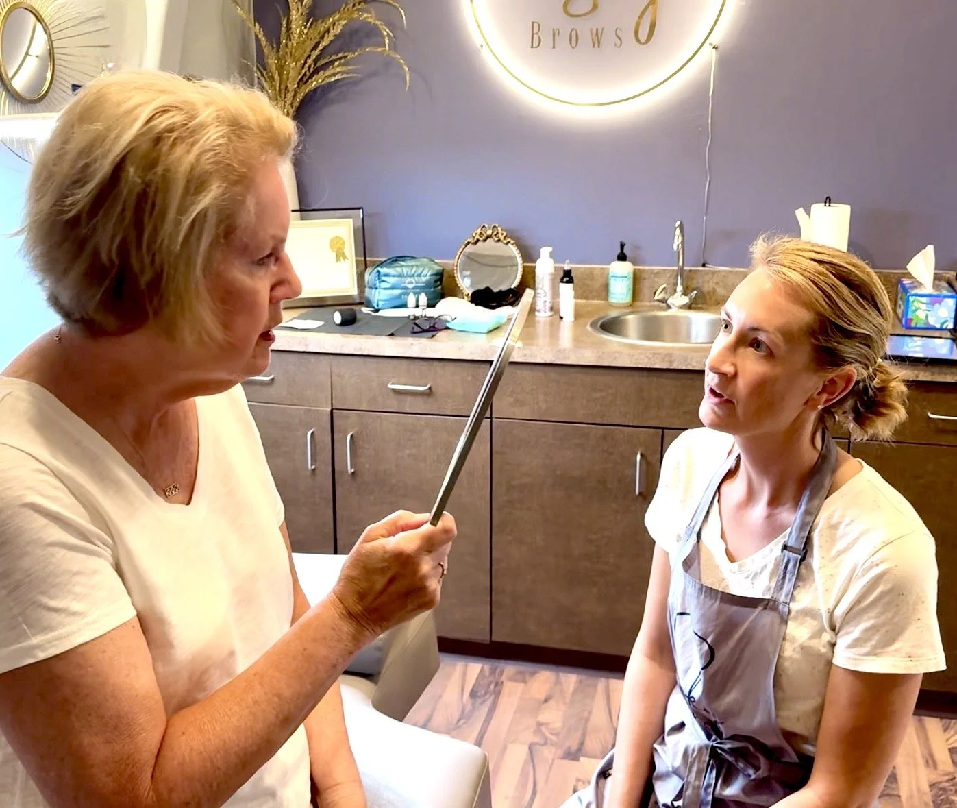 An older woman with short blonde hair holds a pointed stick or tool and appears to be explaining something to a younger woman with her hair tied back, who is sitting on a chair in an examination or treatment room.