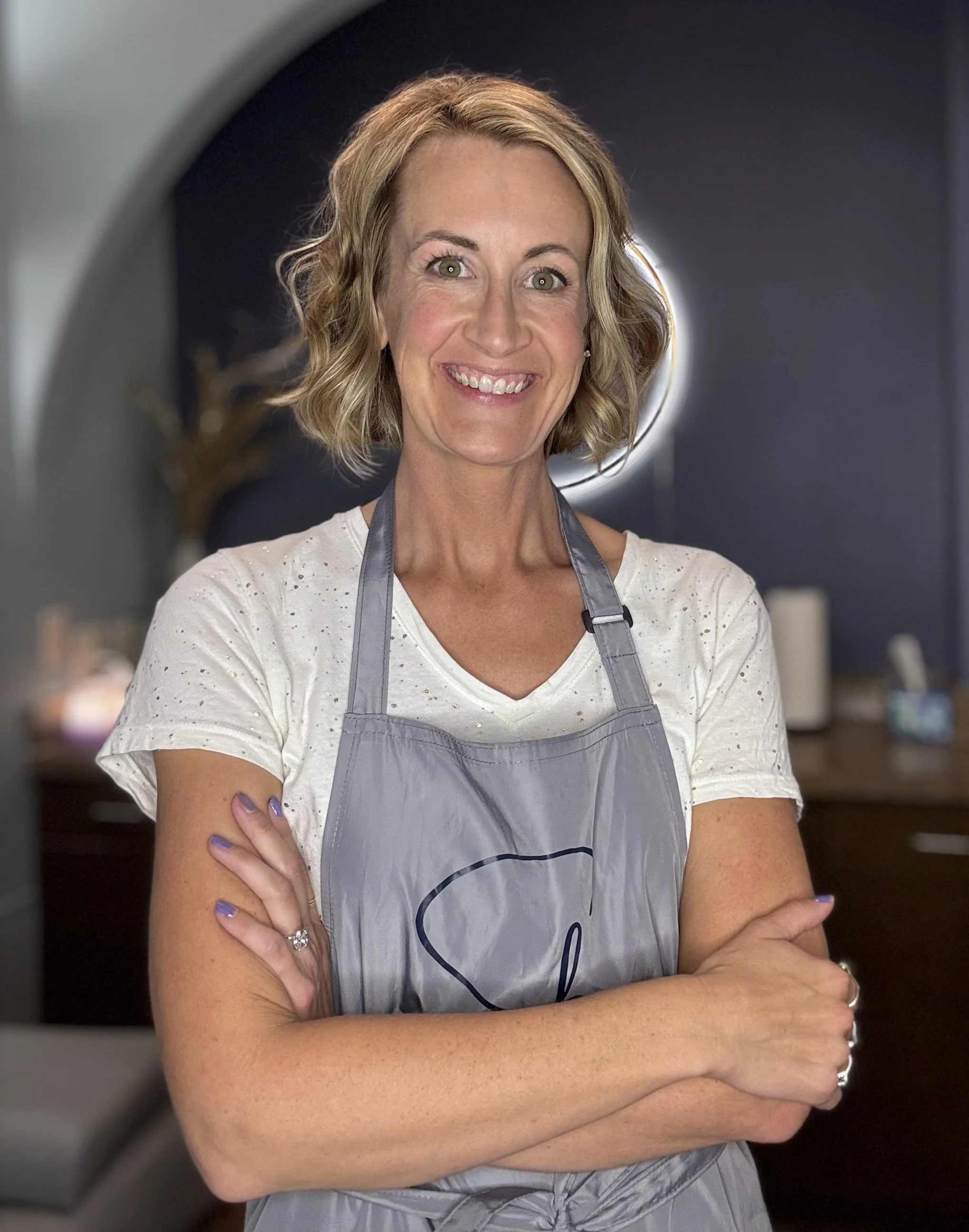 A woman smiling, wearing a light-colored shirt and a gray apron, with crossed arms in front of her. The background shows a circular light behind her head, likely in a kitchen or bakery setting.