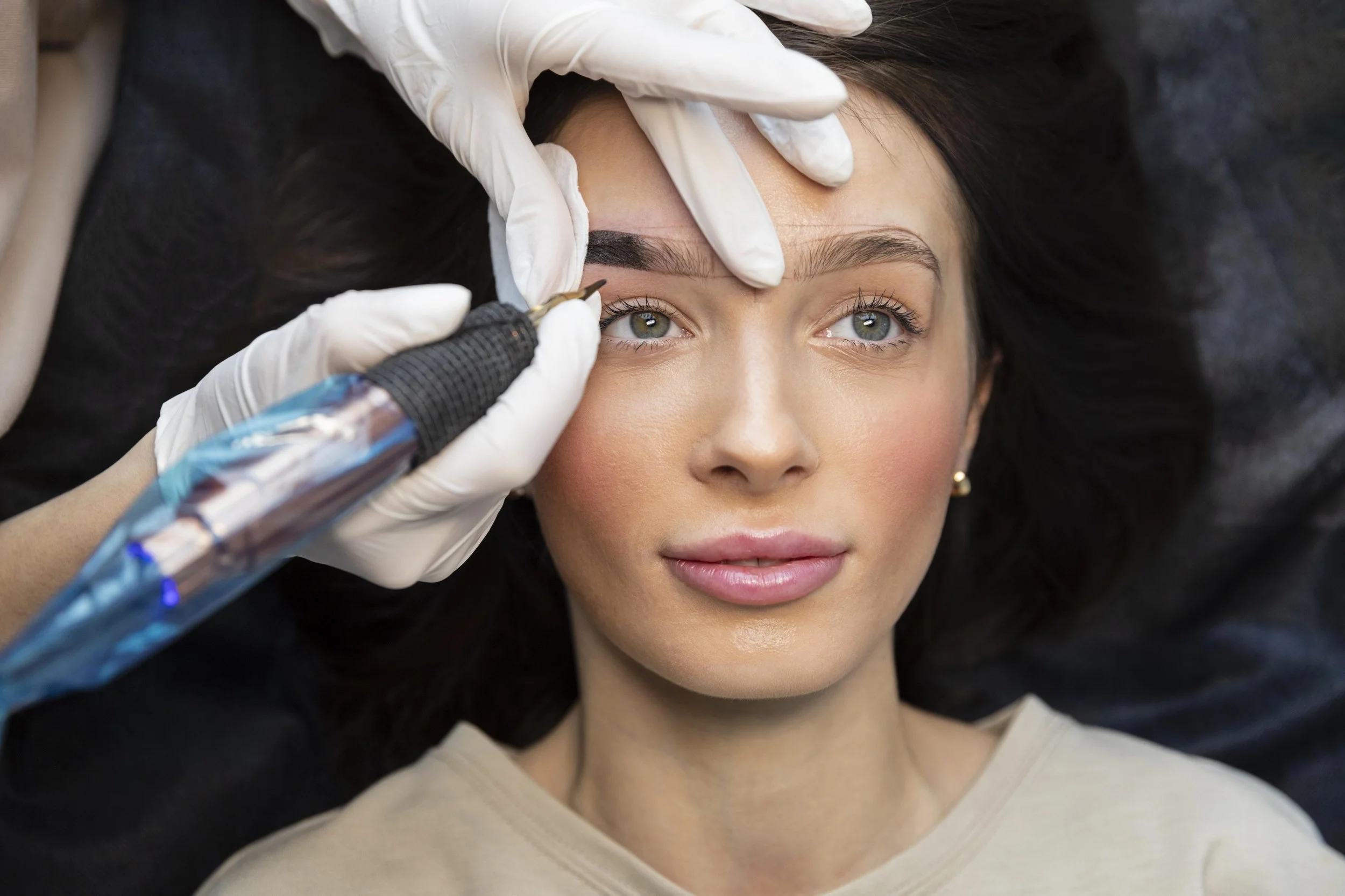 A woman with brown hair and green eyes receiving a cosmetic tattoo procedure on her eyebrows, performed by a professional wearing white gloves.