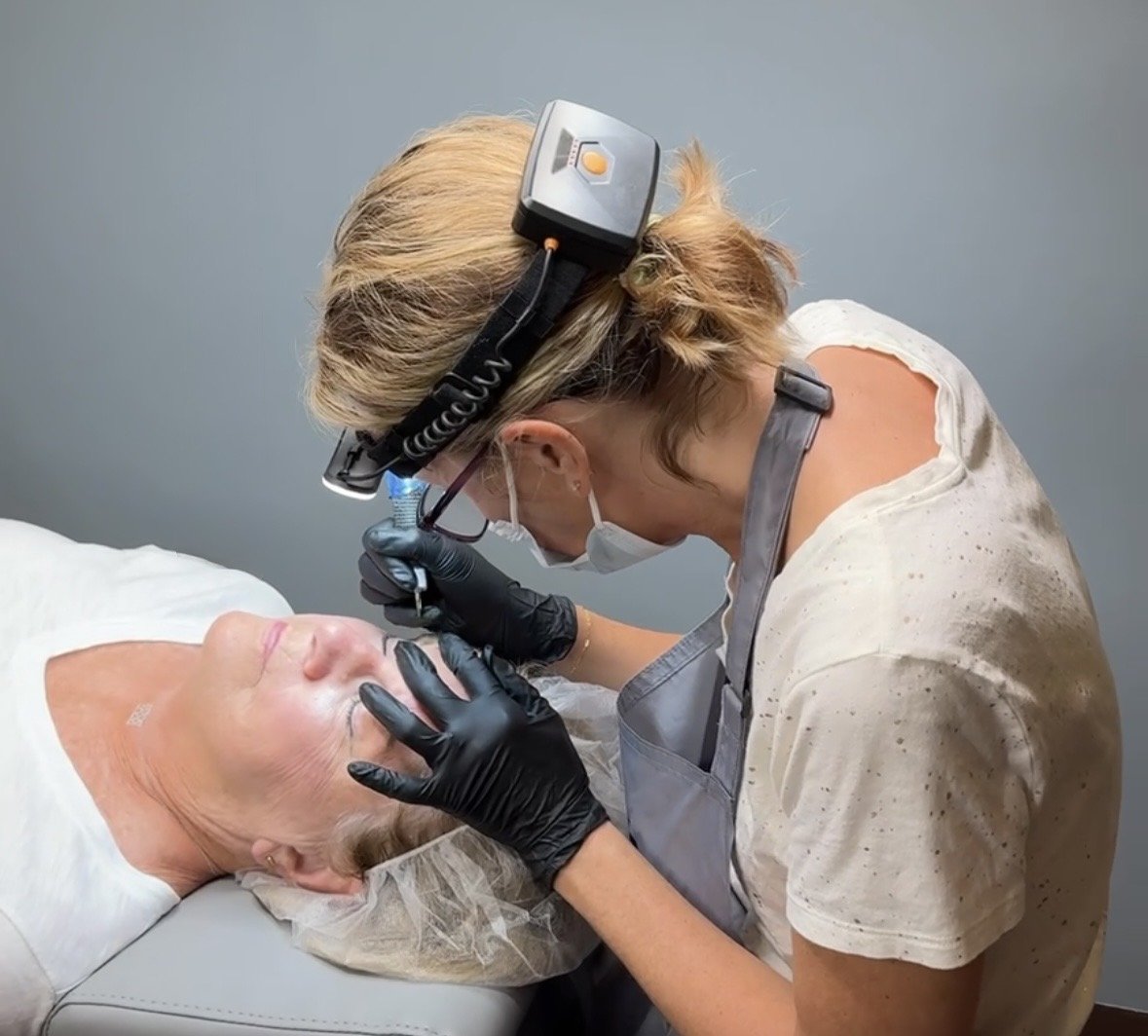 A woman performing a cosmetic procedure on an elderly woman's face, wearing a headlamp, face mask, gloves, and an apron, in a clinical setting.