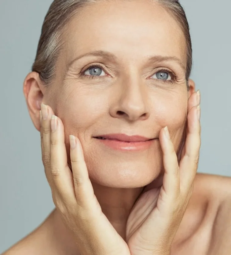 Close-up of a mature woman with clear skin and gray hair, gently touching her face with both hands, smiling lightly, against a neutral background.