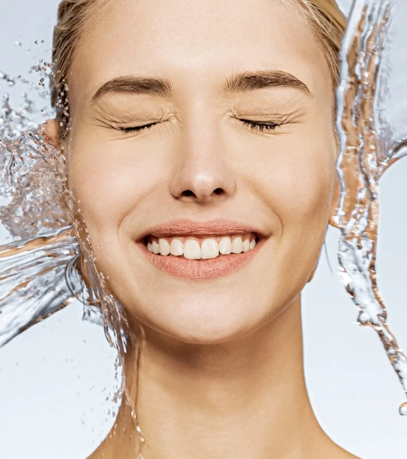 A young woman with closed eyes and a broad smile, rinsing her face with water, with water splashing around her face.