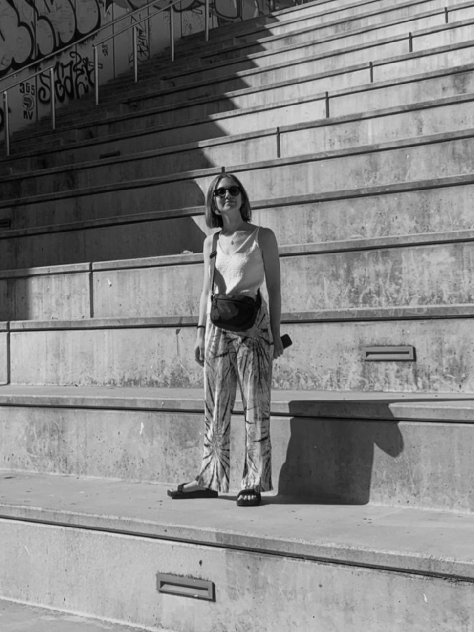 A woman standing on concrete steps with graffiti in the background, wearing sunglasses, a sleeveless top, patterned pants, and sandals, holding a phone in her right hand.